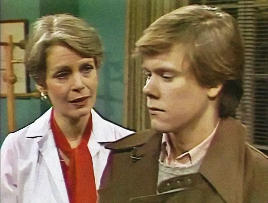 A woman in a white coat and red scarf looks at a young man in a brown coat, who appears serious and is looking away. They are indoors with soft lighting and neutral-colored walls.
