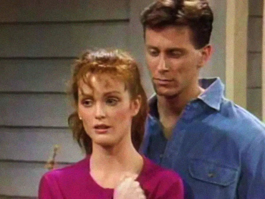 A woman in a pink top stands in front of a man in a blue shirt. They both have serious expressions and are indoors, with a wooden wall in the background.