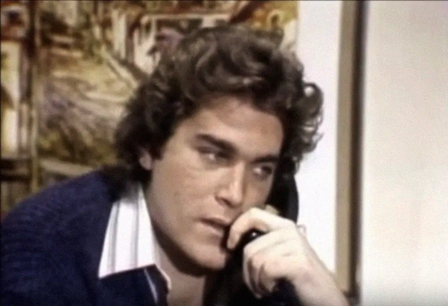 A person with curly brown hair and a thoughtful expression holds their hand to their mouth while looking slightly downward. They wear a dark sweater over a white collared shirt, with a painting visible in the background.