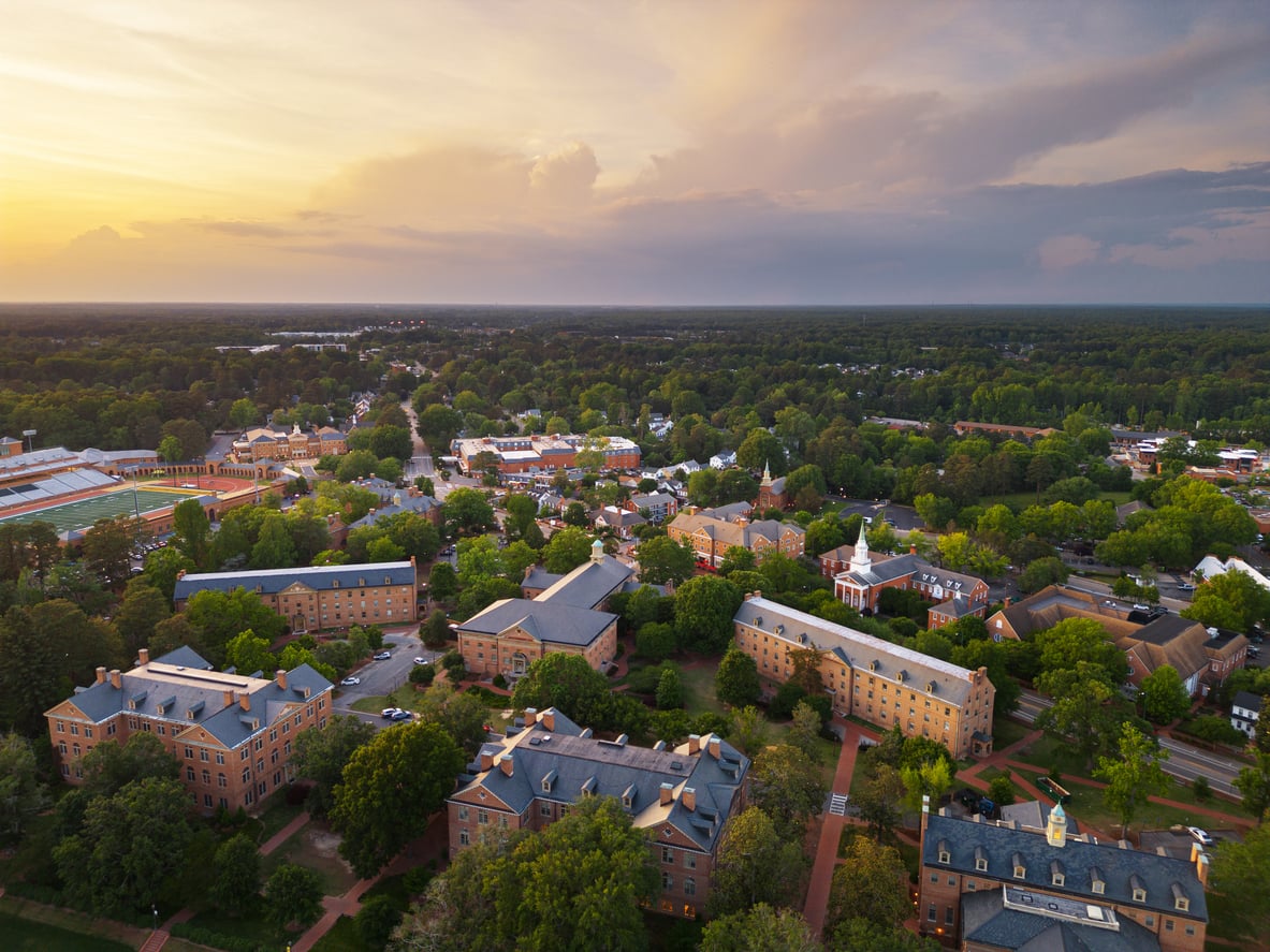 Aerial view of a college campus with brick buildings, green trees, and walkways at sunset. The sky is partly cloudy with soft sunlight, and forested areas surround the campus in the distance—an inviting scene reminiscent of affordable travel destinations that blend natural beauty with vibrant academic life.