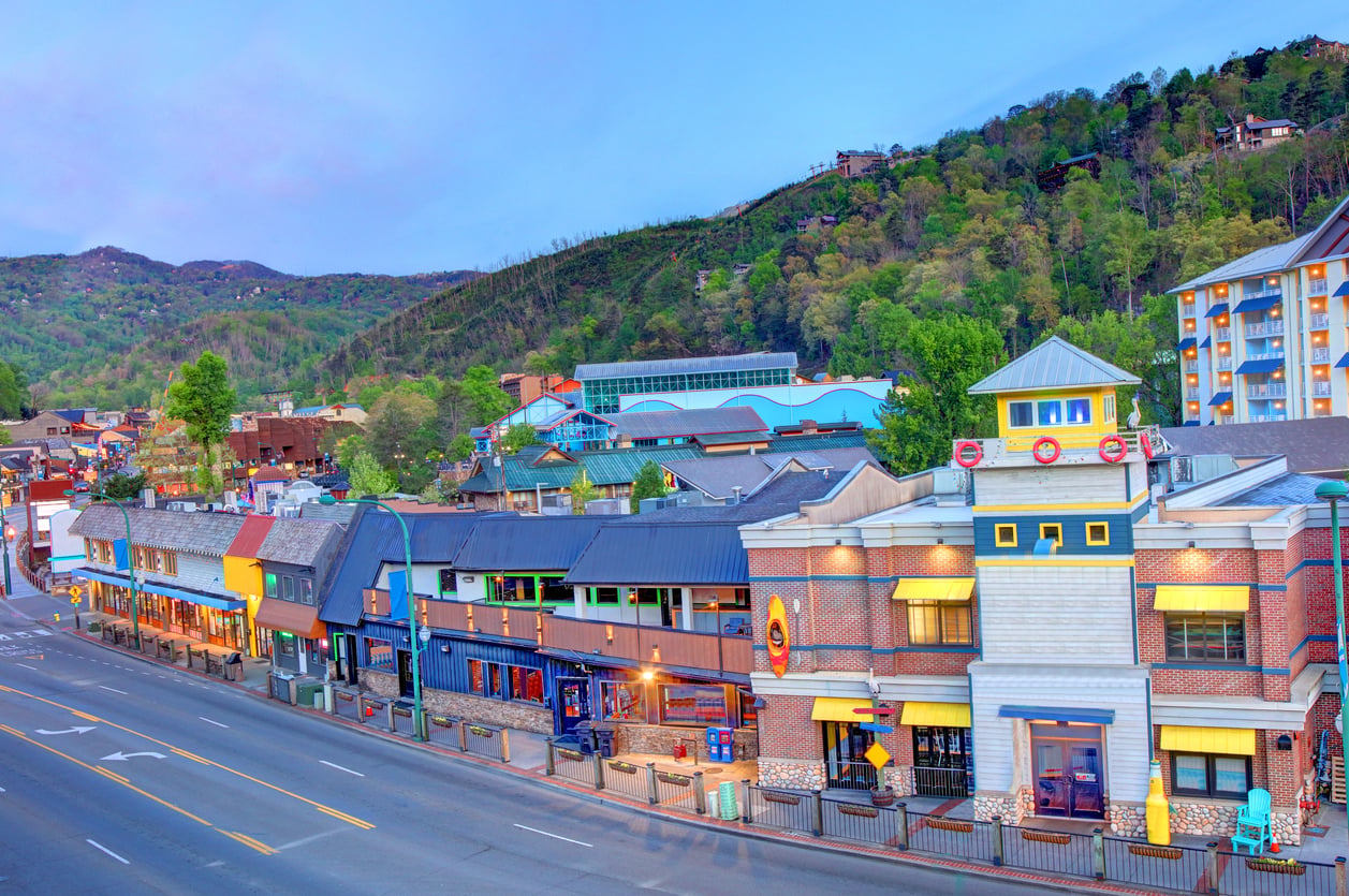 Colorful shops and restaurants line a quiet street at dusk, set against a backdrop of green, tree-covered hills and a partly cloudy sky—showcasing one of the region’s most charming and affordable travel destinations.