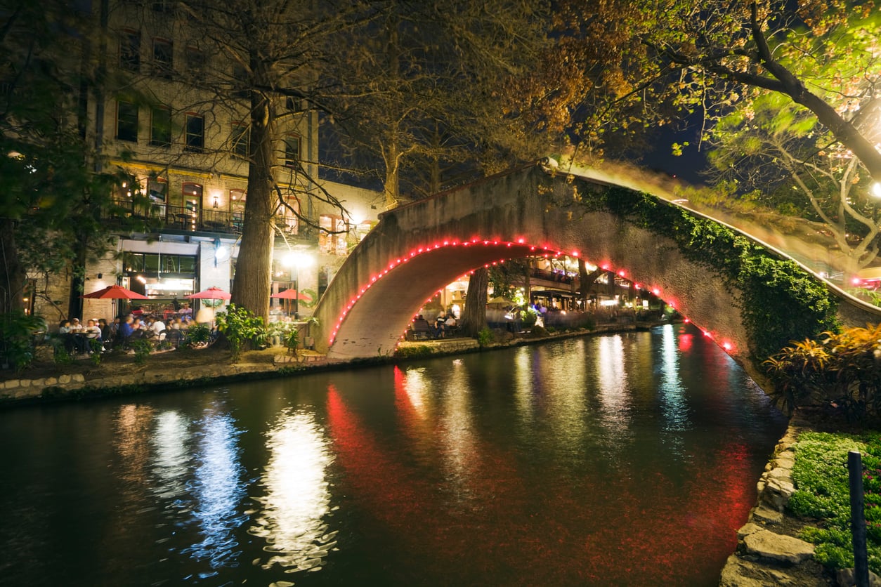 A stone bridge with red lights arches over a calm river at night, reflecting the lights. People dine by the riverside under umbrellas, surrounded by trees and illuminated buildings—one of the region's most picturesque and affordable travel destinations.