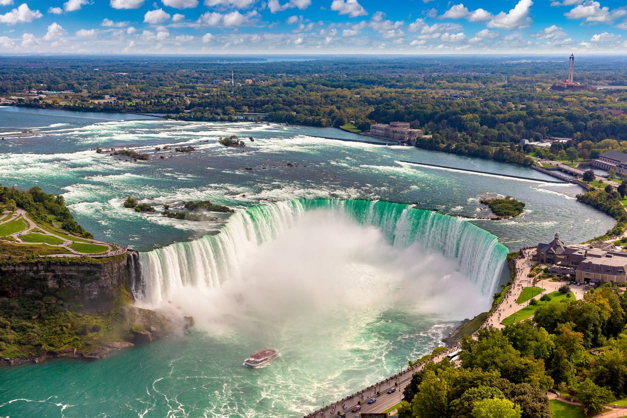 Aerial view of Niagara Falls, one of the most affordable travel destinations, with a tour boat near the base, mist rising from the waterfall, lush greenery, and buildings in the background under a partly cloudy sky.