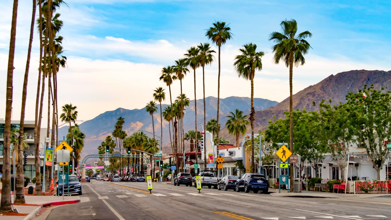 A street lined with palm trees, parked cars, and shops—typical of many affordable travel destinations—stretches ahead, with mountains in the background under a partly cloudy sky. Pedestrian crossings and street signs are visible.