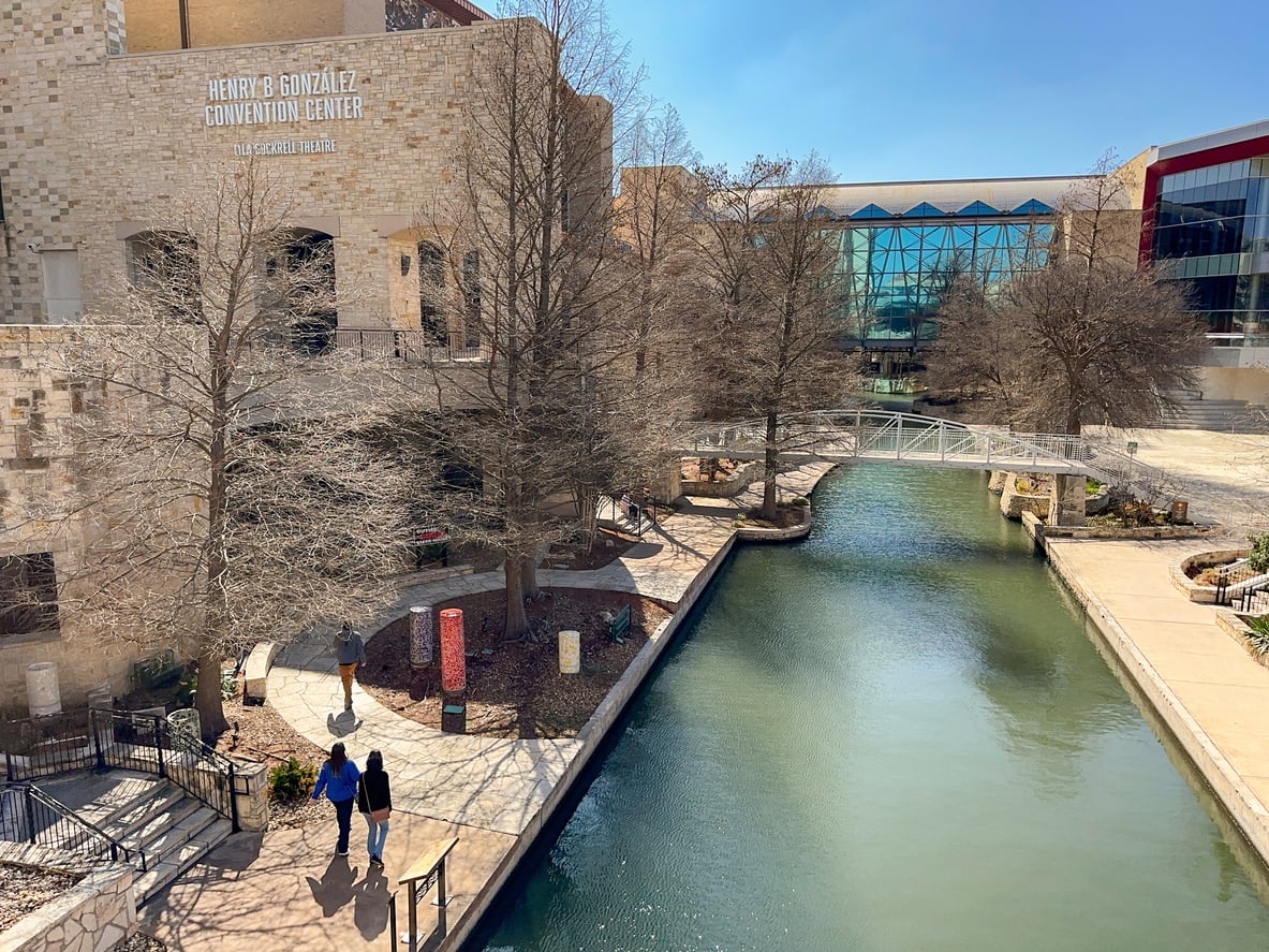 Two people walk along a path beside a canal near the Henry B. González Convention Center, with leafless trees, a small bridge, and modern buildings in the background under a clear blue sky—capturing the charm of one of the region’s affordable travel destinations.