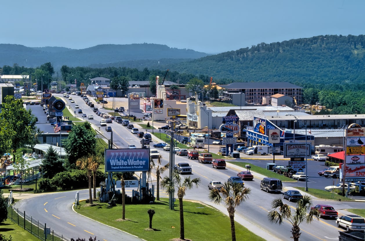 A busy roadway lined with shops, restaurants, and billboards, offering a glimpse into one of the area's most affordable travel destinations. Cars drive in both directions, while green, wooded hills and clear skies form a picturesque backdrop.