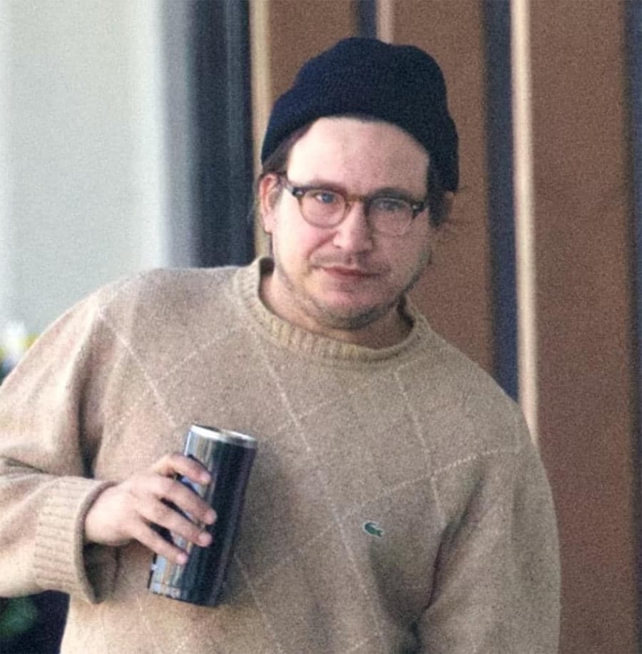 A person wearing glasses, a dark beanie, and a beige sweater holds a tumbler cup while standing indoors.
