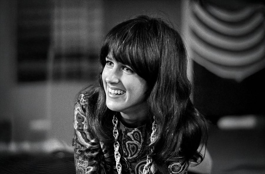 A woman with long dark hair and bangs smiles while wearing a paisley-patterned top and a chunky necklace. The background is softly blurred with geometric patterns.