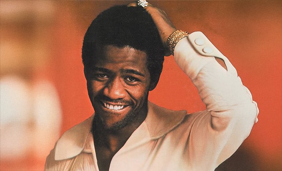 A smiling man in a light shirt stands against a warm, orange background, touching the back of his head with one hand. He wears a gold ring and bracelet, and has short, curly hair.