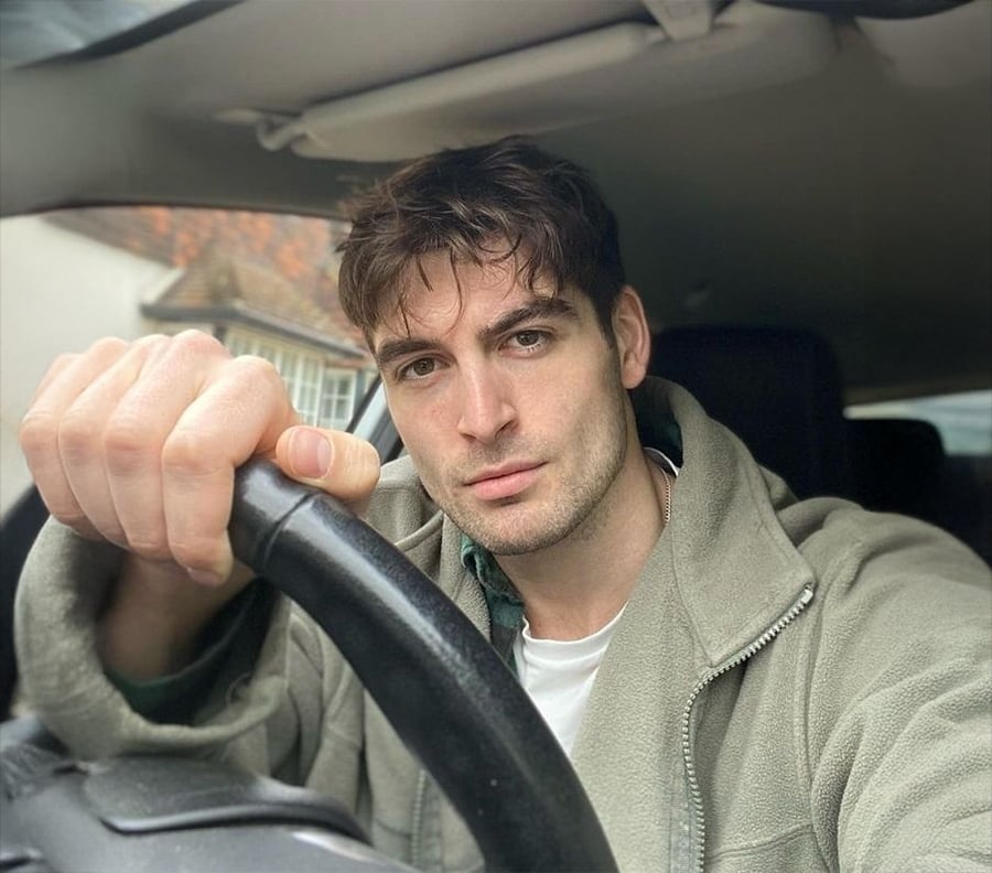 A man with tousled brown hair sits in a car, holding the steering wheel with one hand and looking at the camera. He wears a light grey zip-up jacket over a white shirt.