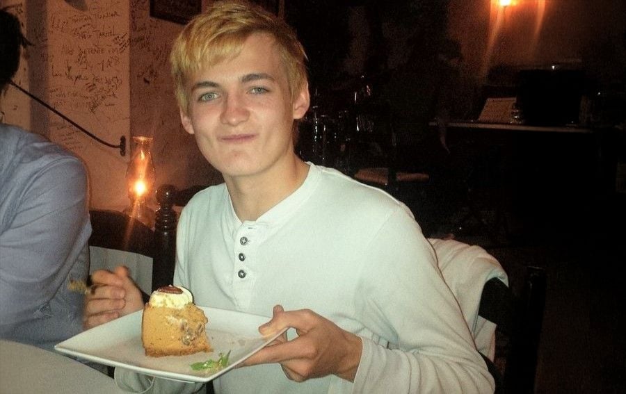 Un joven de cabello rubio, vestido con camisa blanca, sonríe mientras sostiene un plato con un trozo de pastel y un tenedor, sentado en un restaurante con poca luz.