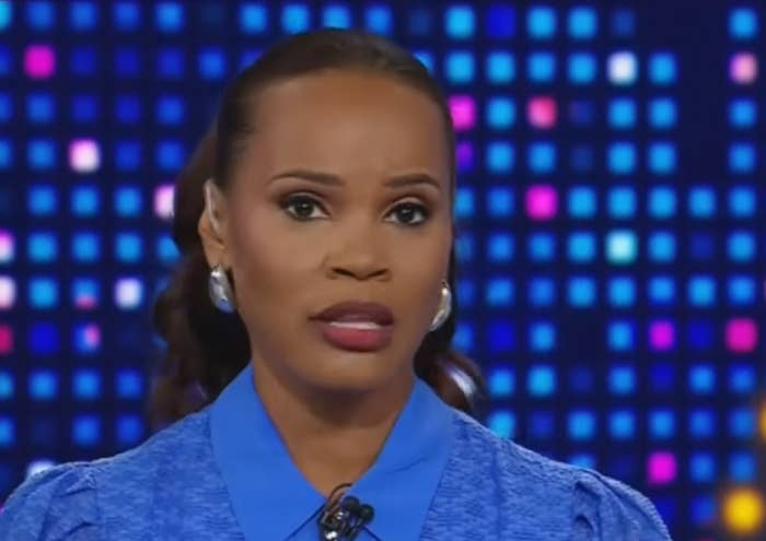 A news anchor delivers a broadcast, wearing a formal blue top with silver earrings. A digital display with a pixel pattern is in the background