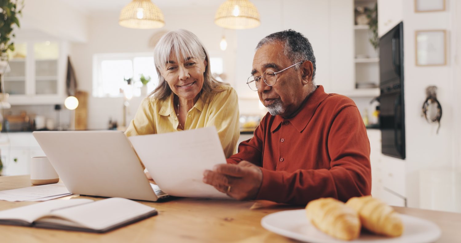 A senior woman with light grey hair and a yellow collared shirt smiles while looking at papers held by a senior man with grey hair, a beard, and glasses, wearing a rust-colored collared shirt. They are seated at a wooden table with a white laptop, a white mug, and an open notebook. In the foreground, two croissants rest on a white plate. The background shows a brightly lit, modern kitchen interior with light wood and white cabinetry.