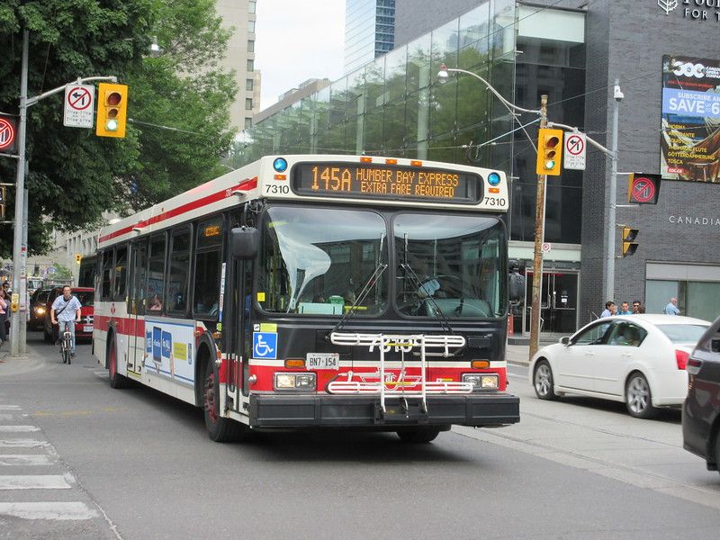 Un autobus Humber Bay Express 145A de la Toronto Transit Commission attend à une intersection de la ville. Un cycliste passe à côté, et des voitures et des piétons se trouvent à proximité. Des immeubles de grande hauteur et des feux de signalisation sont visibles en arrière-plan.
