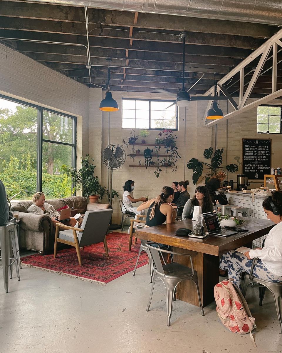 Les gens travaillent et socialisent dans un café confortable et ensoleillé avec des plantes, de grandes fenêtres et des plafonds apparents. Certains s'assoient à des tables avec des ordinateurs portables, tandis que d'autres se détendent sur des canapés près d'un tapis rouge. L'atmosphère est décontractée et accueillante.