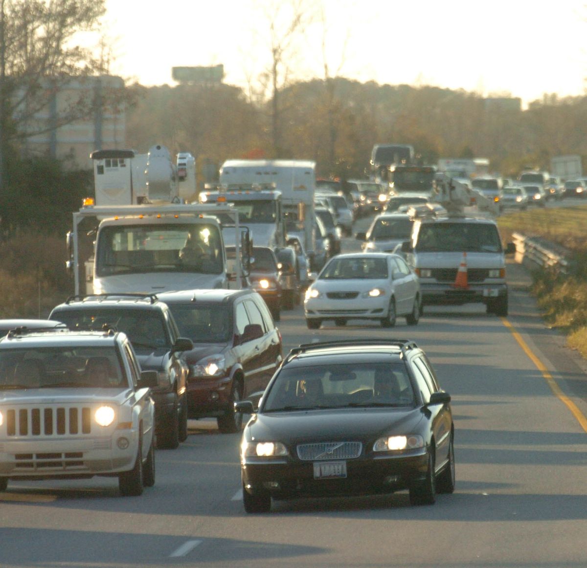 Une circulation dense de voitures et de camions occupe plusieurs voies sur une autoroute pendant la journée, certains véhicules étant arrêtés et d'autres avançant lentement. Des arbres et une barrière bordent la route.