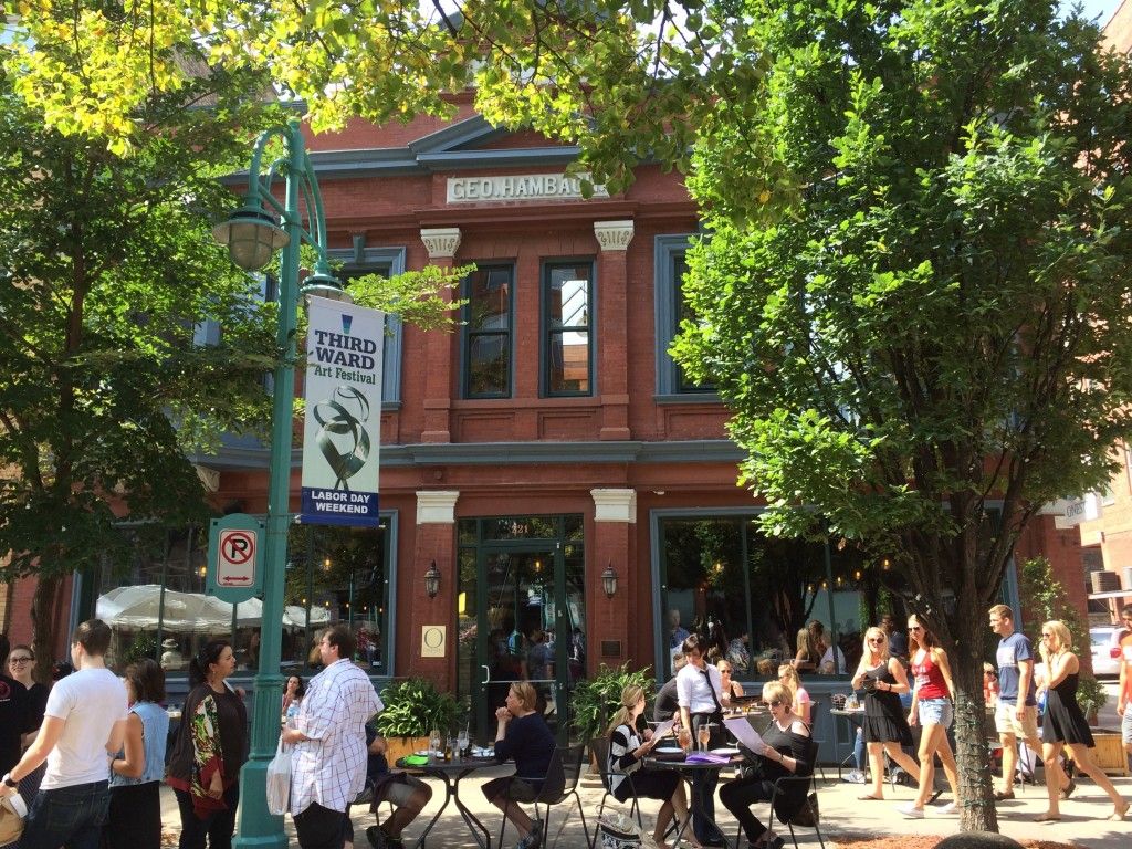 Les gens se rassemblent devant un bâtiment en briques rouges doté de grandes fenêtres, d’arbres et de tables extérieures. Un panneau annonce le Third Ward Art Festival le week-end de la fête du Travail. La lumière du soleil filtre à travers les arbres, créant une scène de rue animée.