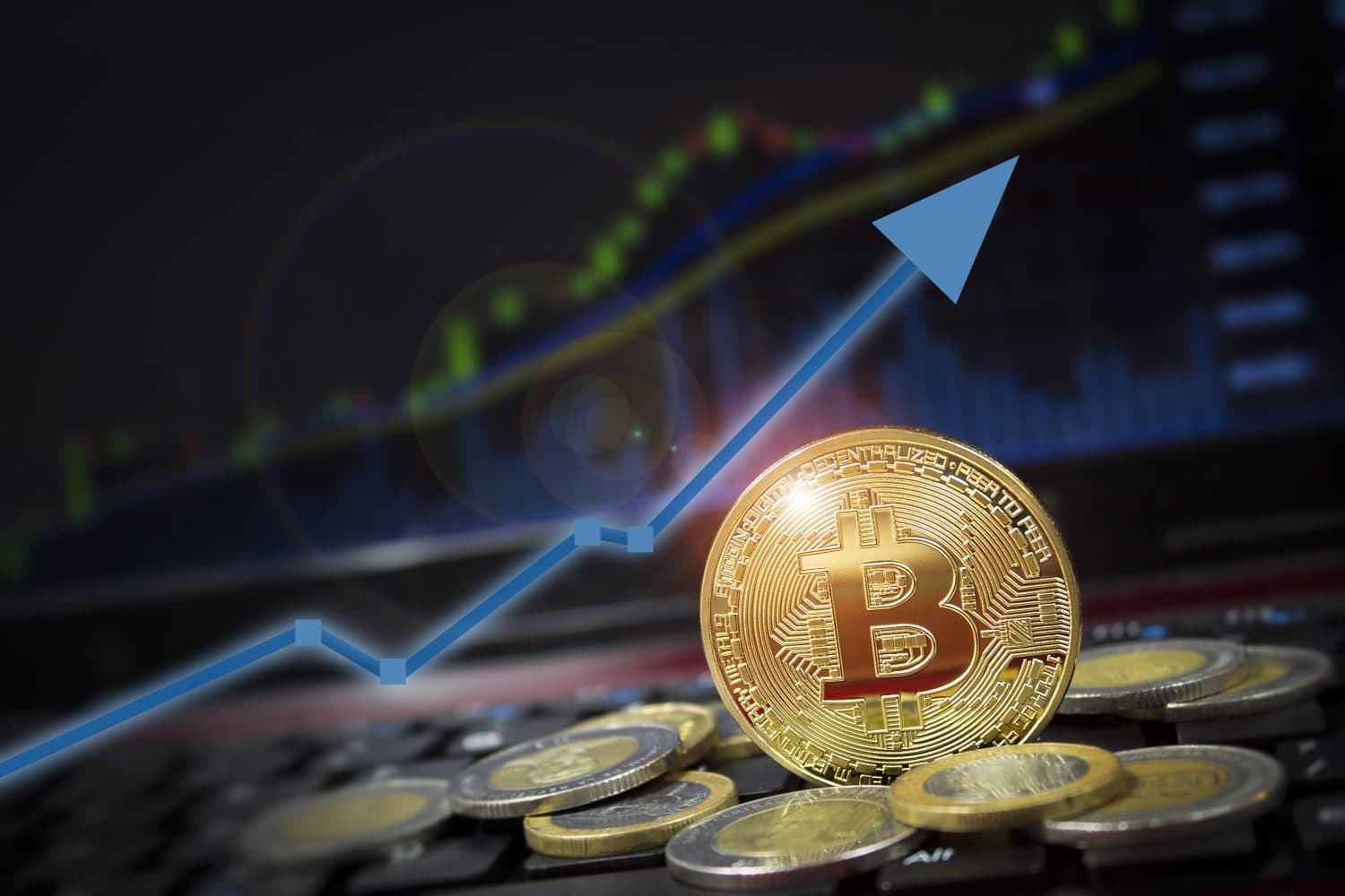 A golden Bitcoin coin stands prominently on a dark keyboard, surrounded by various silver and gold-toned physical coins. In the blurred background, a digital stock chart on a screen displays an upward trend, highlighted by a bright blue line with an arrow pointing towards the upper right corner. The scene is illuminated by a soft lens flare.