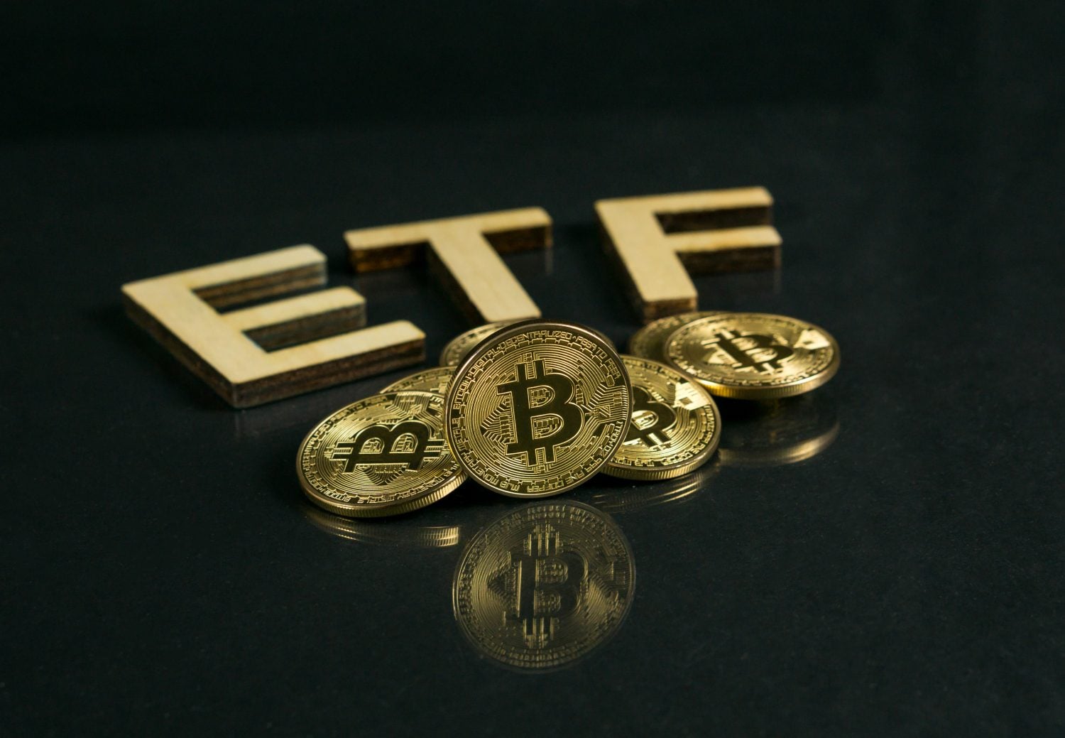 Golden Bitcoin coins are scattered in front of three wooden block letters spelling 'ETF' on a dark, reflective surface. The coins reflect on the surface below, creating a sense of depth and focus on the central Bitcoin coin displaying its 'B' logo.