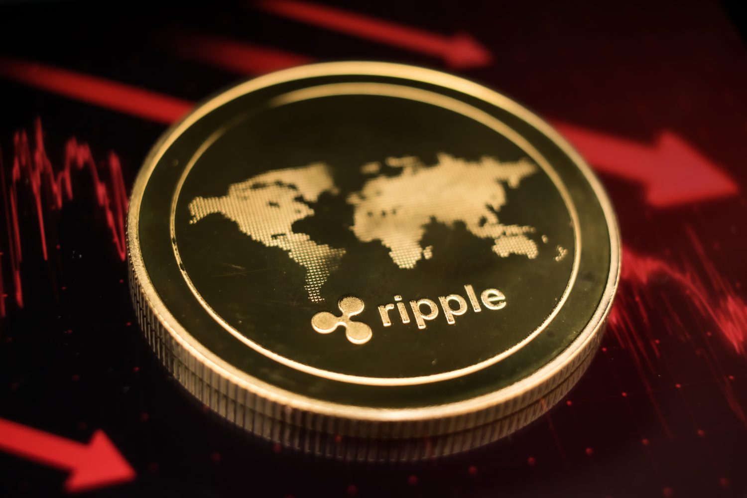 A close-up shot of a golden Ripple (XRP) cryptocurrency coin, resting on its edge. The coin's face displays a world map design and the 'ripple' logo with text. The background is dark, illuminated by glowing red lines forming a downward trend chart and large red arrows pointing downwards, indicating a market decline.