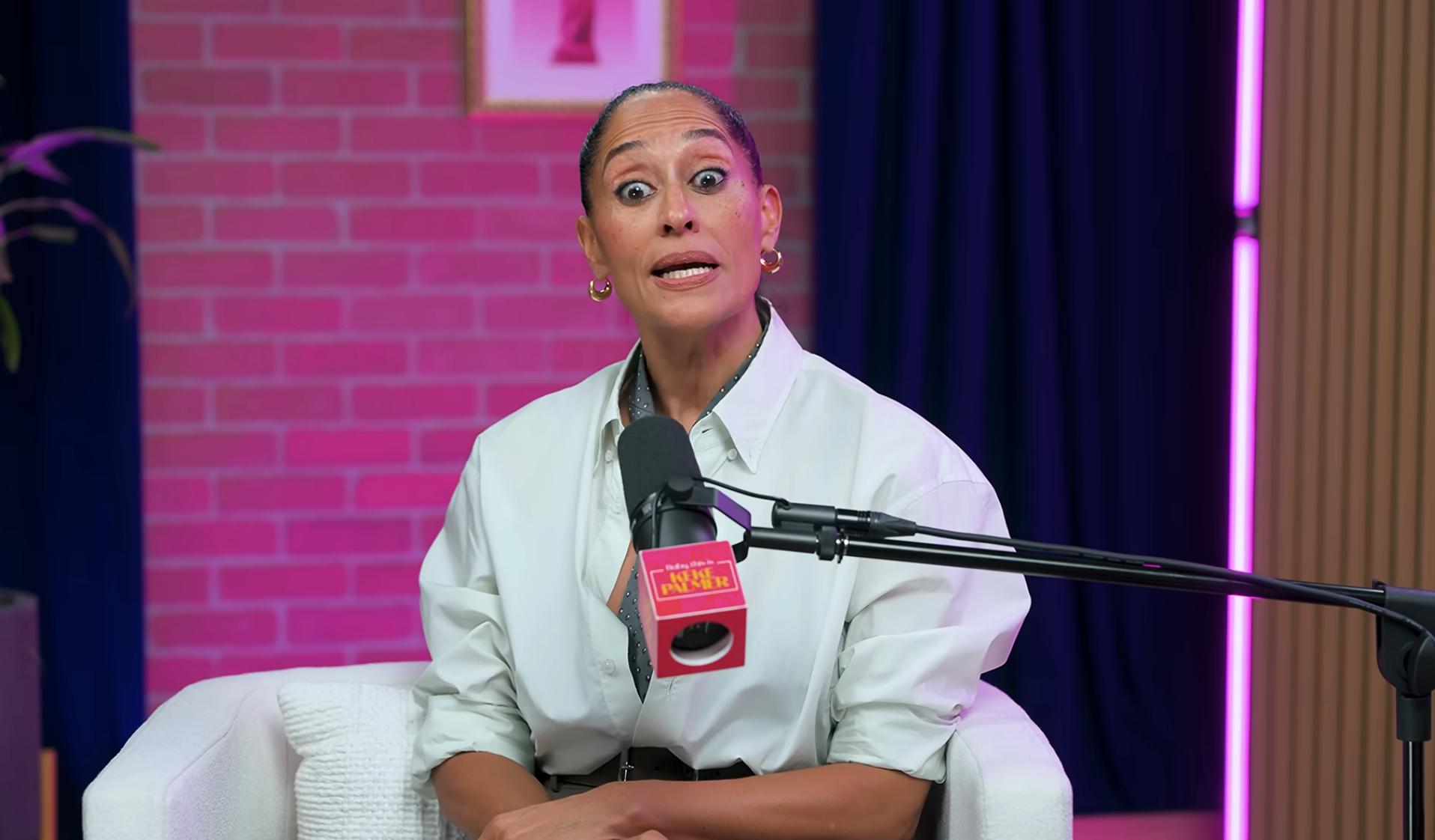 Una persona con un elegante top blanco se sienta en un estudio de podcast y habla animadamente por un micrófono
