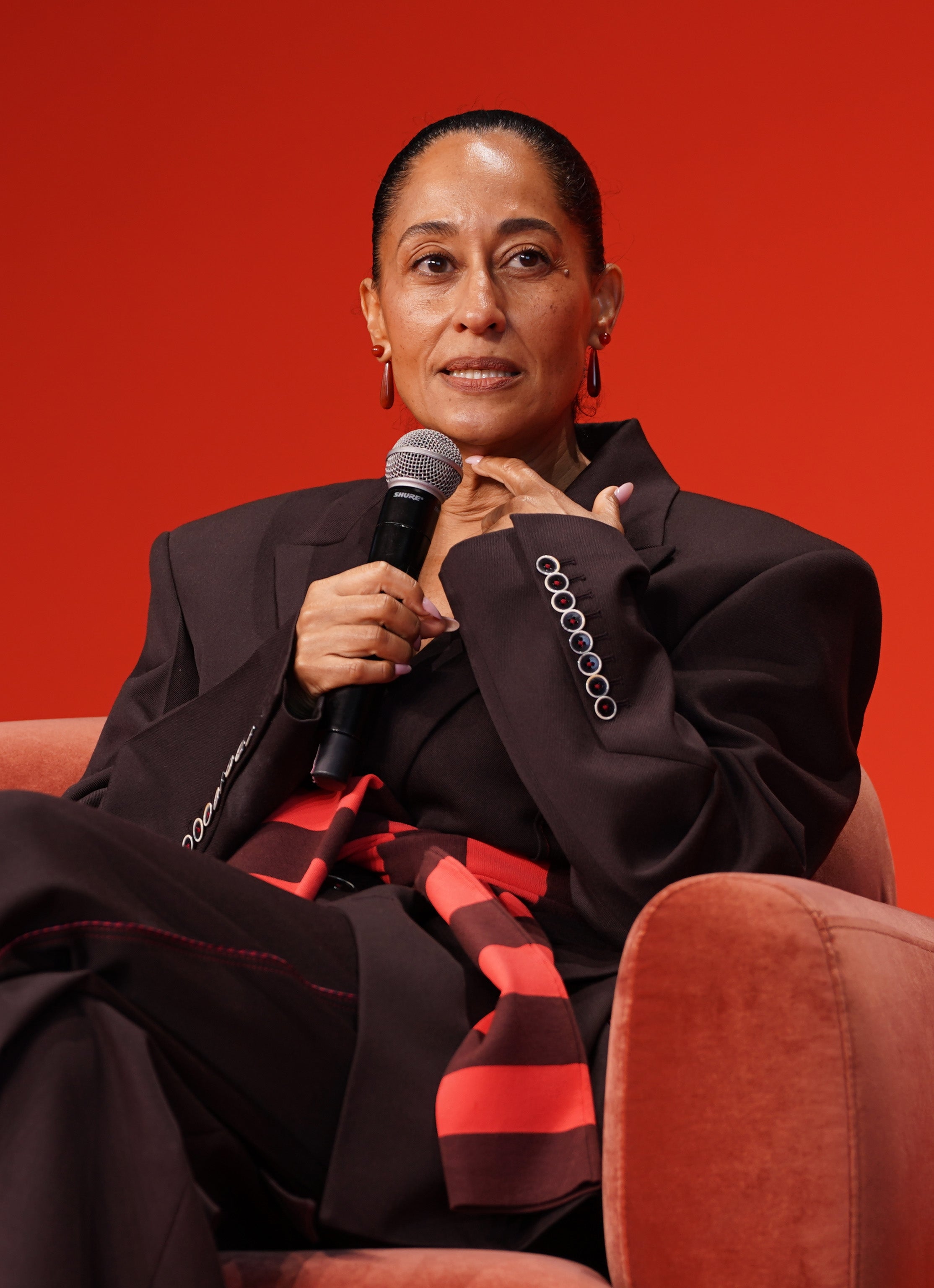 Persona con un elegante traje oscuro de gran tamaño con detalles en rojo, sosteniendo un micrófono mientras está sentada en un entorno de entrevista