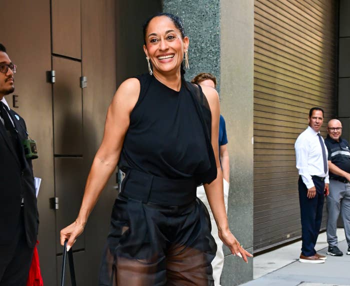 Persona con una blusa negra sin mangas y pantalones elegantes, sonriendo mientras camina afuera, con otras personas al fondo