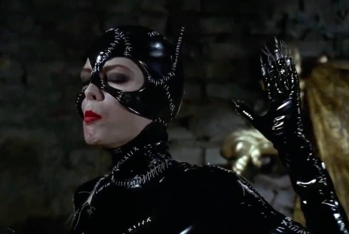 Pessoa com macacão de couro costurado e máscara, imitando pose felina; lembra um personagem clássico de filme