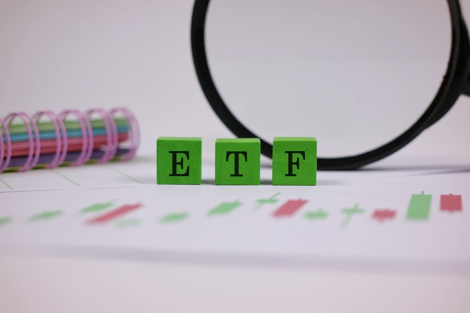 Three green wooden blocks with black letters spell 'ETF' in the foreground. They rest on a white document showing a blurred financial chart with red and green candlesticks. A large magnifying glass with a black rim is visible in the upper right background, and a colorful spiral-bound notebook is blurred in the upper left background.