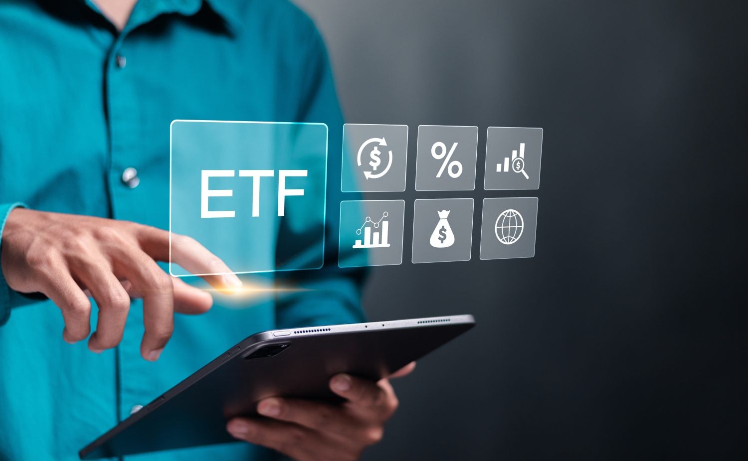 A person wearing a teal-colored long-sleeved shirt holds a dark tablet with their left hand, while their right index finger points towards a glowing, translucent rectangular digital interface. The interface displays the white text 'ETF' prominently, next to a grid of six smaller translucent squares containing white financial icons: a dollar sign with refresh arrows, a percentage sign, a bar chart with a magnifying glass, a line graph, a money bag with a dollar sign, and a globe. A bright golden light emanates from the point where the finger touches the 'ETF' box. The background is a dark, blurred gray, suggesting a modern, digital environment.