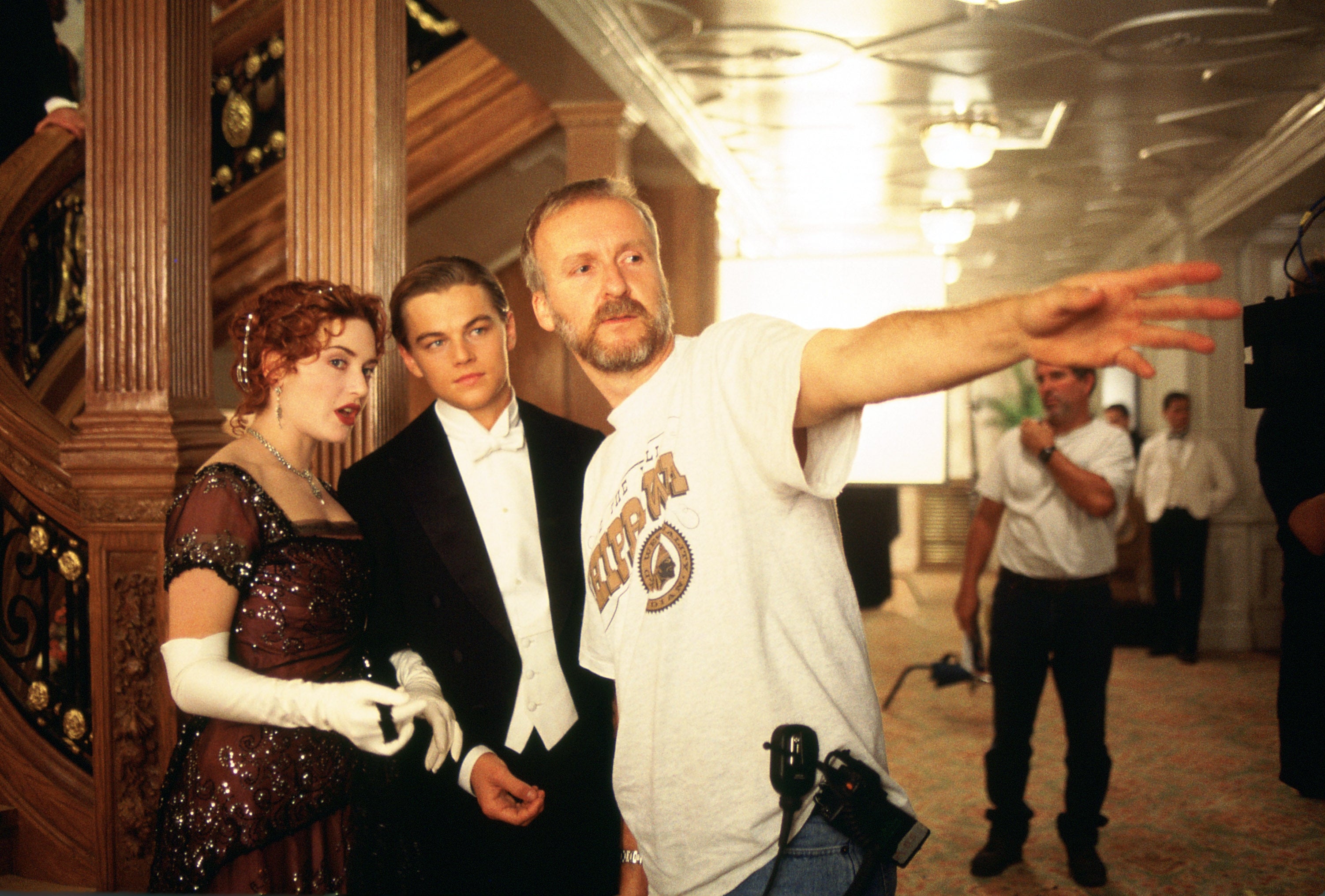 Actors in period attire on a film set with a director gesturing, likely discussing a scene. The setting is reminiscent of an ornate ship interior