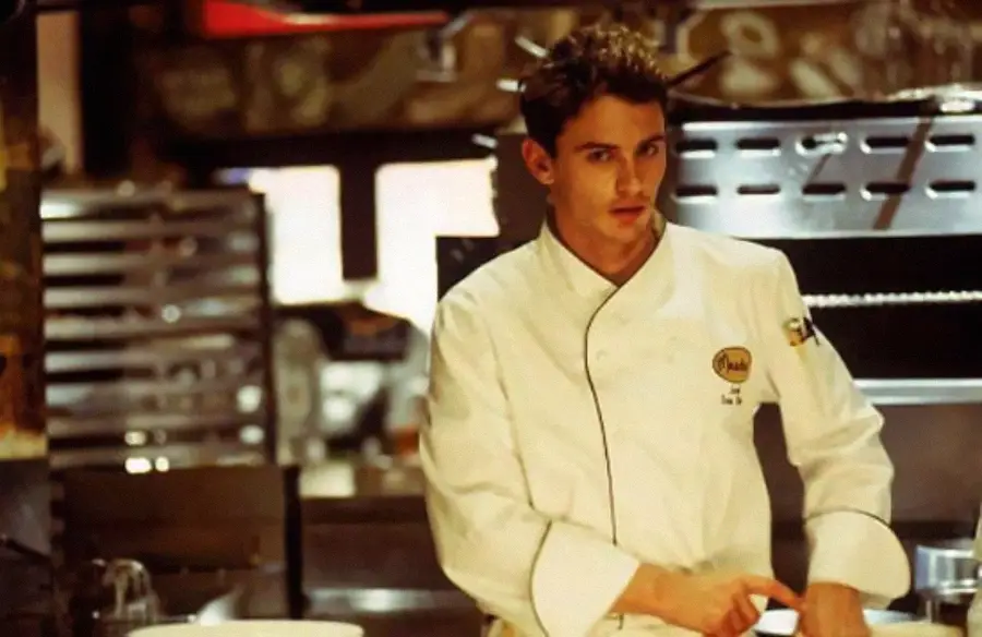 A young chef in a white jacket stands in a busy kitchen, looking at the camera while preparing food. Stainless steel equipment and trays are visible in the background.