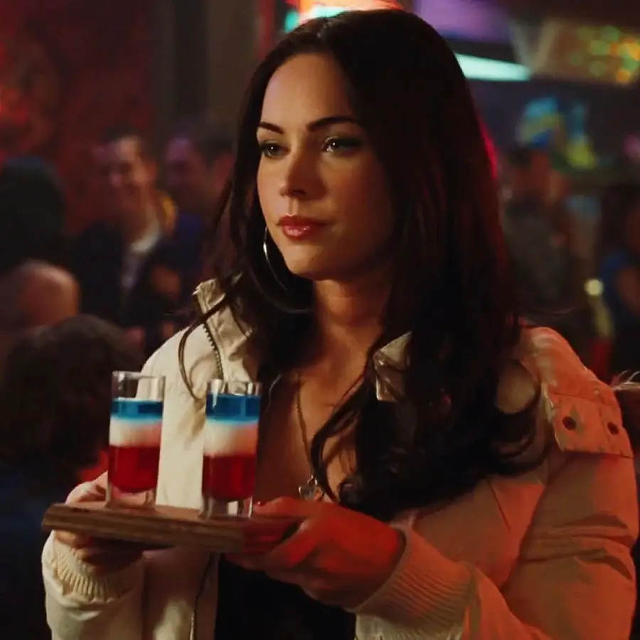 A woman with long dark hair, wearing a white jacket, holds a wooden tray with two layered red, white, and blue shot drinks at a lively bar or club.