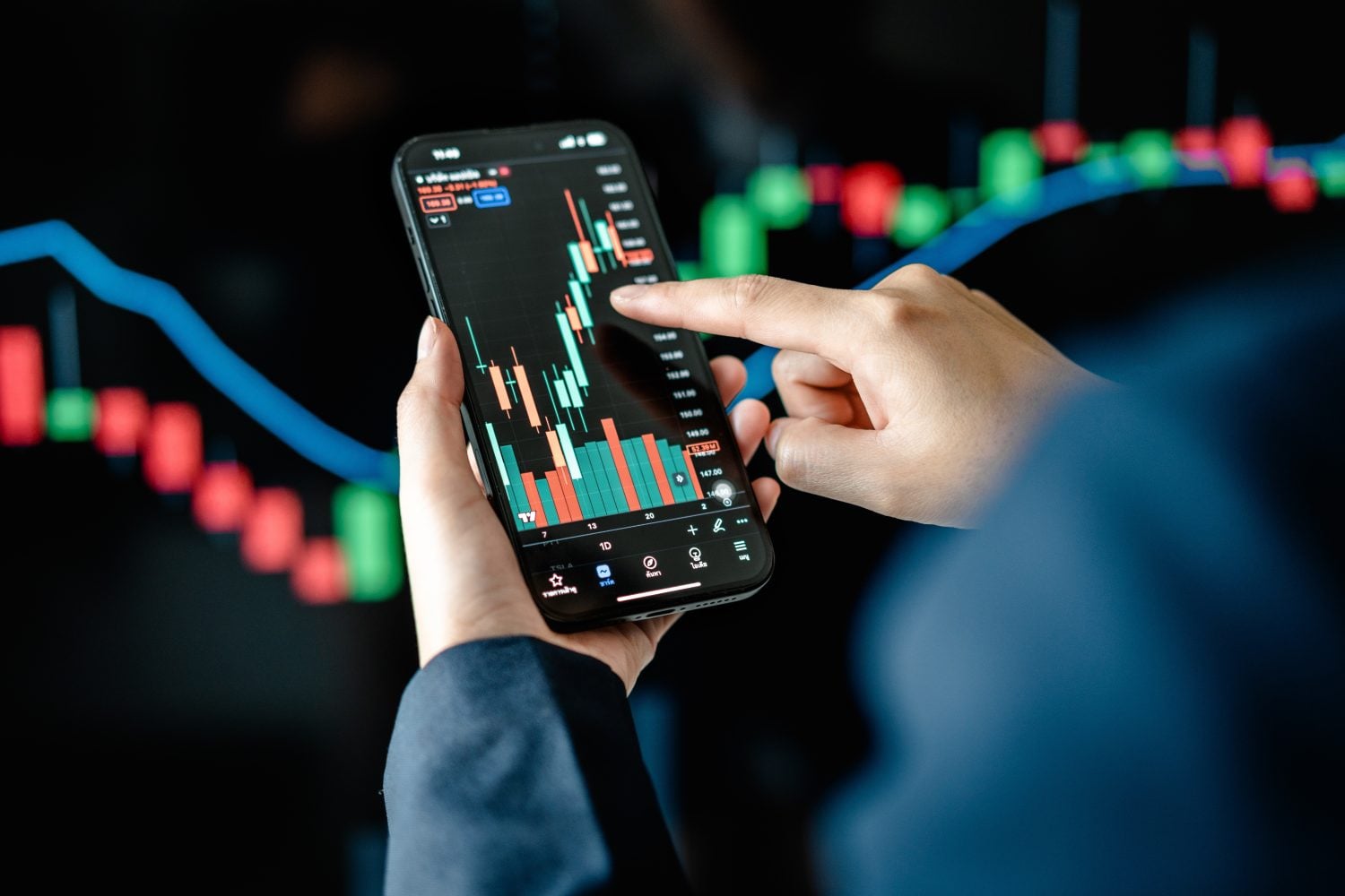A close-up shot shows a person holding a black smartphone horizontally, displaying a vibrant green and red candlestick stock chart against a dark background. A finger from the right hand is pointing to a specific point on the chart. In the blurred background, another larger screen displays a similar financial chart with a prominent blue trend line and red and green vertical bars.