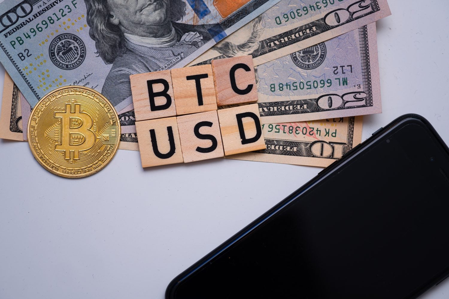 A top-down view of financial items on a white background. A golden Bitcoin coin is prominently displayed on the left. Scattered US dollar banknotes, including $100 and $50 bills, are beneath and around six wooden letter tiles arranged to spell 'BTC' on the top row and 'USD' on the bottom row. A black smartphone is partially visible in the bottom right corner.