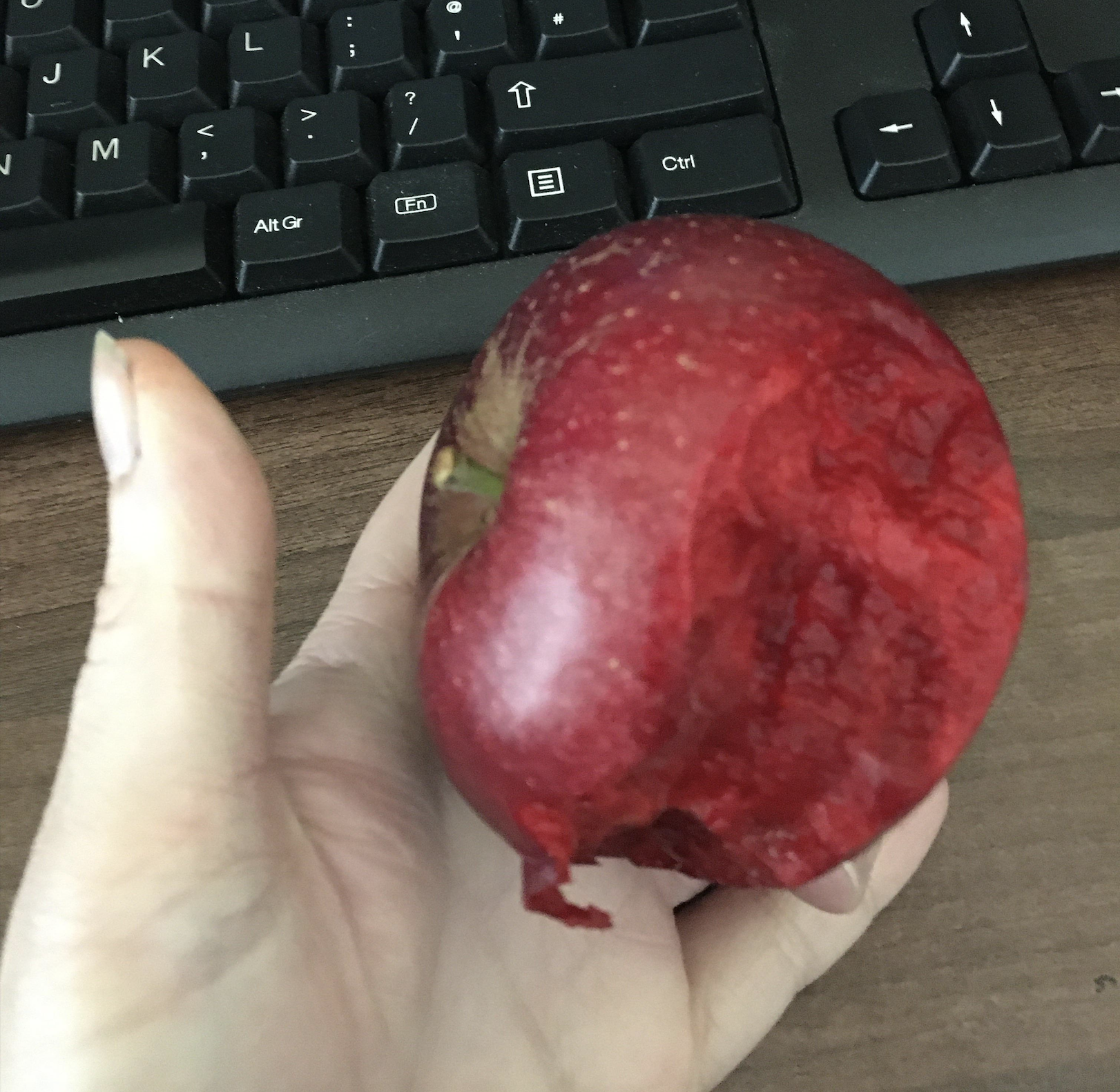 A hand holding a red apple with a large bite taken out of it, in front of a black computer keyboard on a wooden desk.