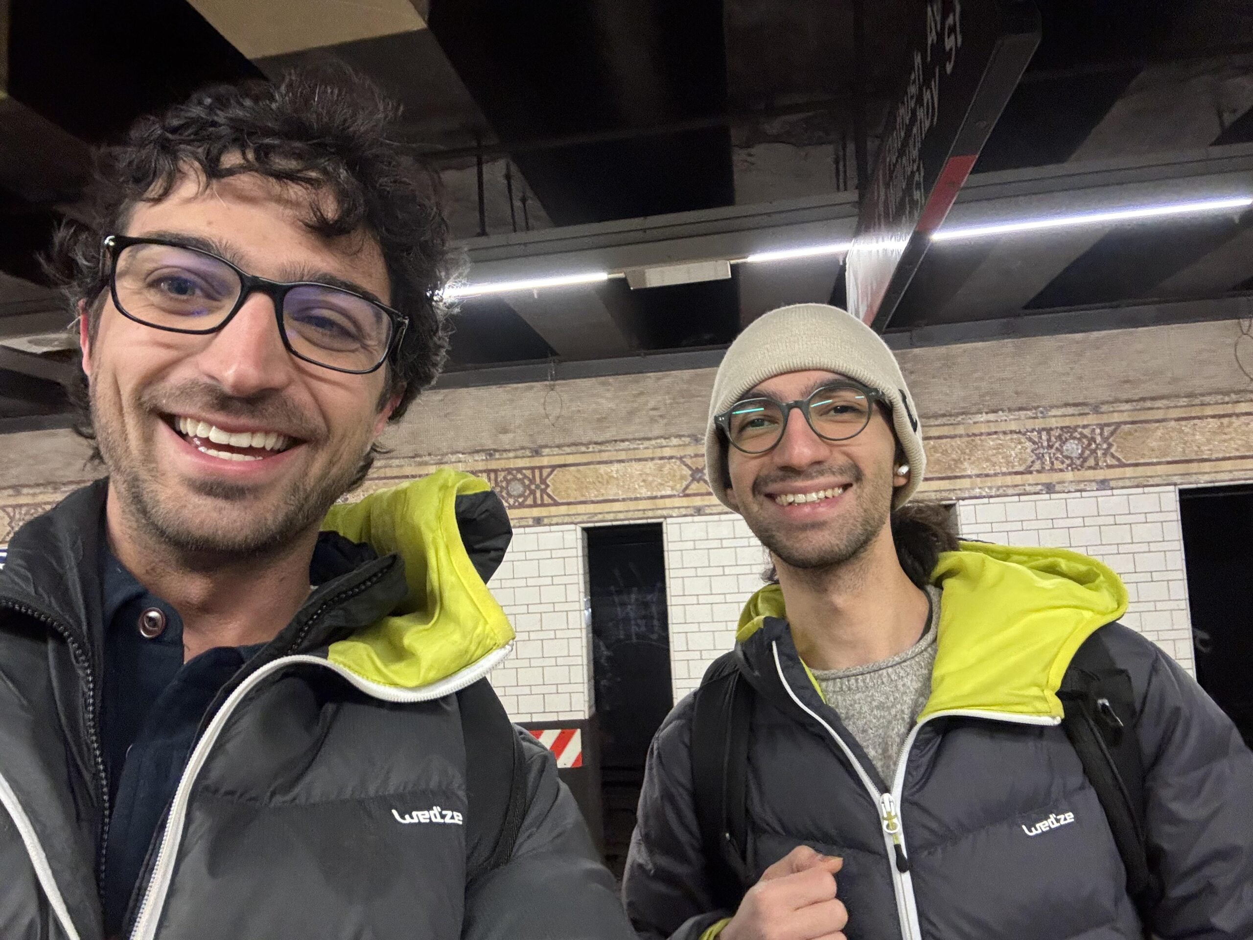 Two people wearing glasses and matching jackets with yellow-green linings smile at the camera while standing on a subway platform with white tiled walls in the background.
