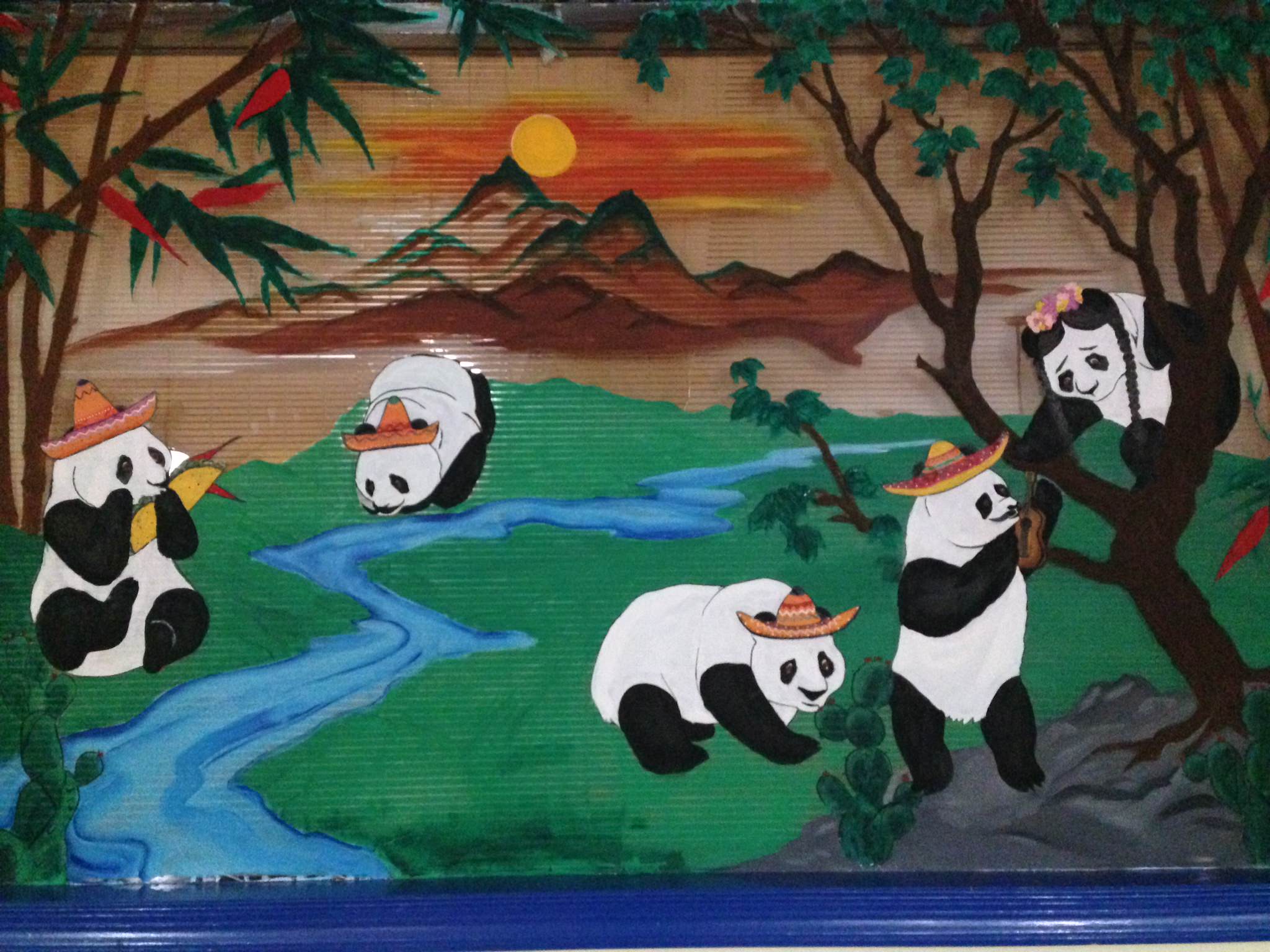 Five pandas wearing hats play and eat in a grassy landscape, with a river, bamboo, mountains, and a sunset in the background. One panda is picking fruit from a tree.