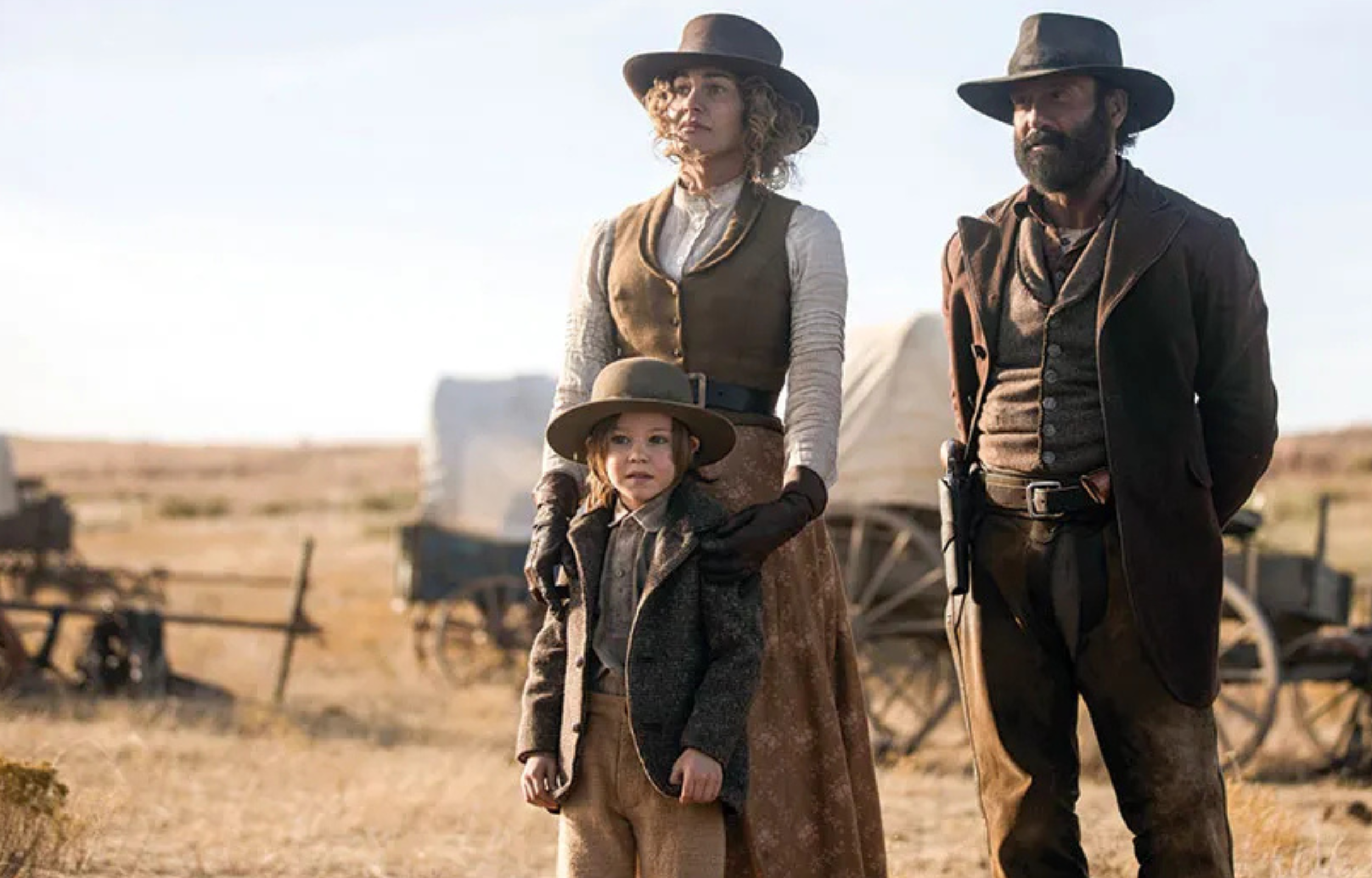 Three individuals, including a child, dressed in western attire, standing in an open landscape with wagons in the background.