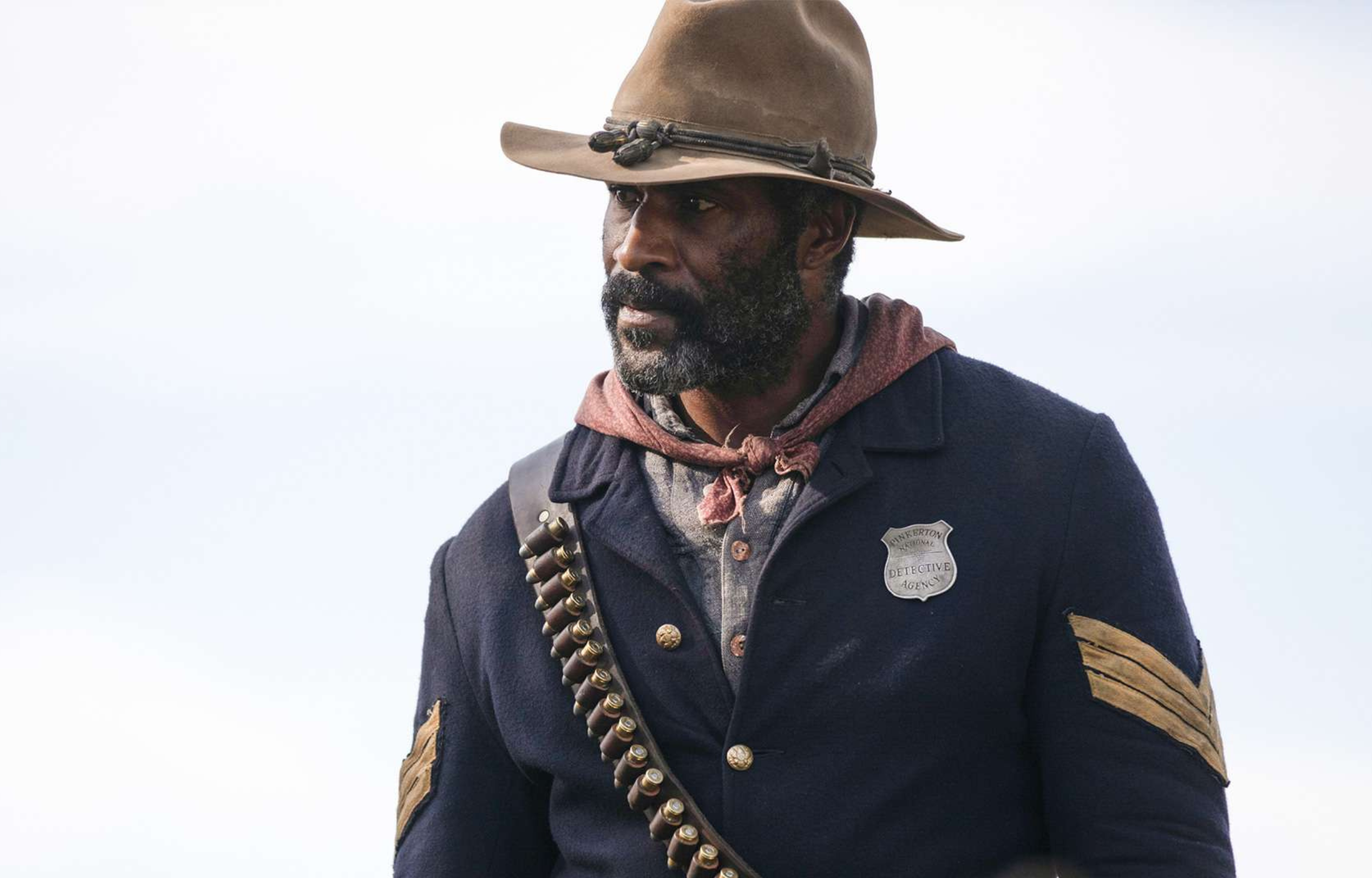Man in a historical uniform with a badge, hat, and scarf, standing outdoors against a cloudy sky.