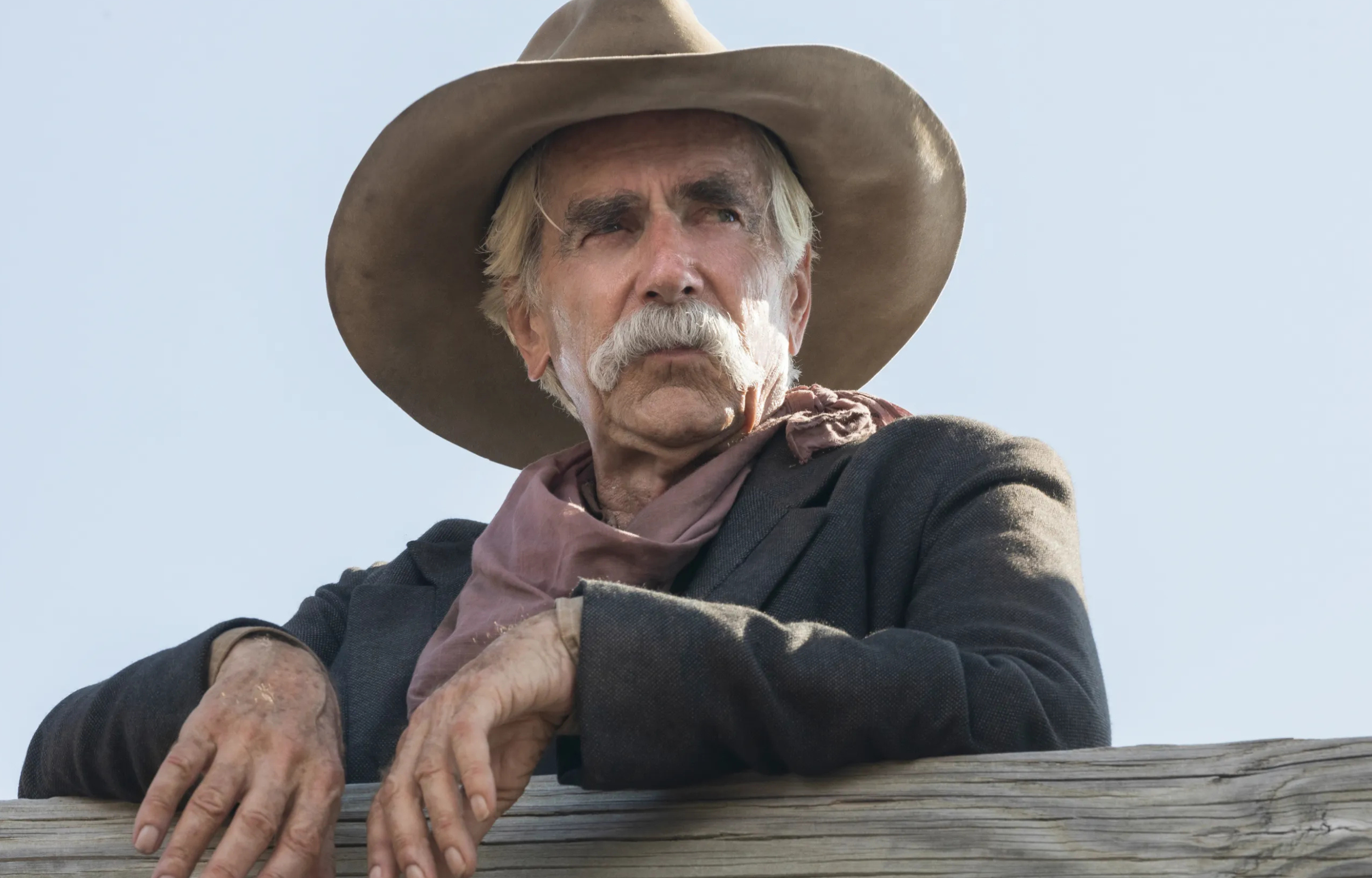 Elderly man wearing a cowboy hat and scarf, leaning against a wooden railing with a serious expression.