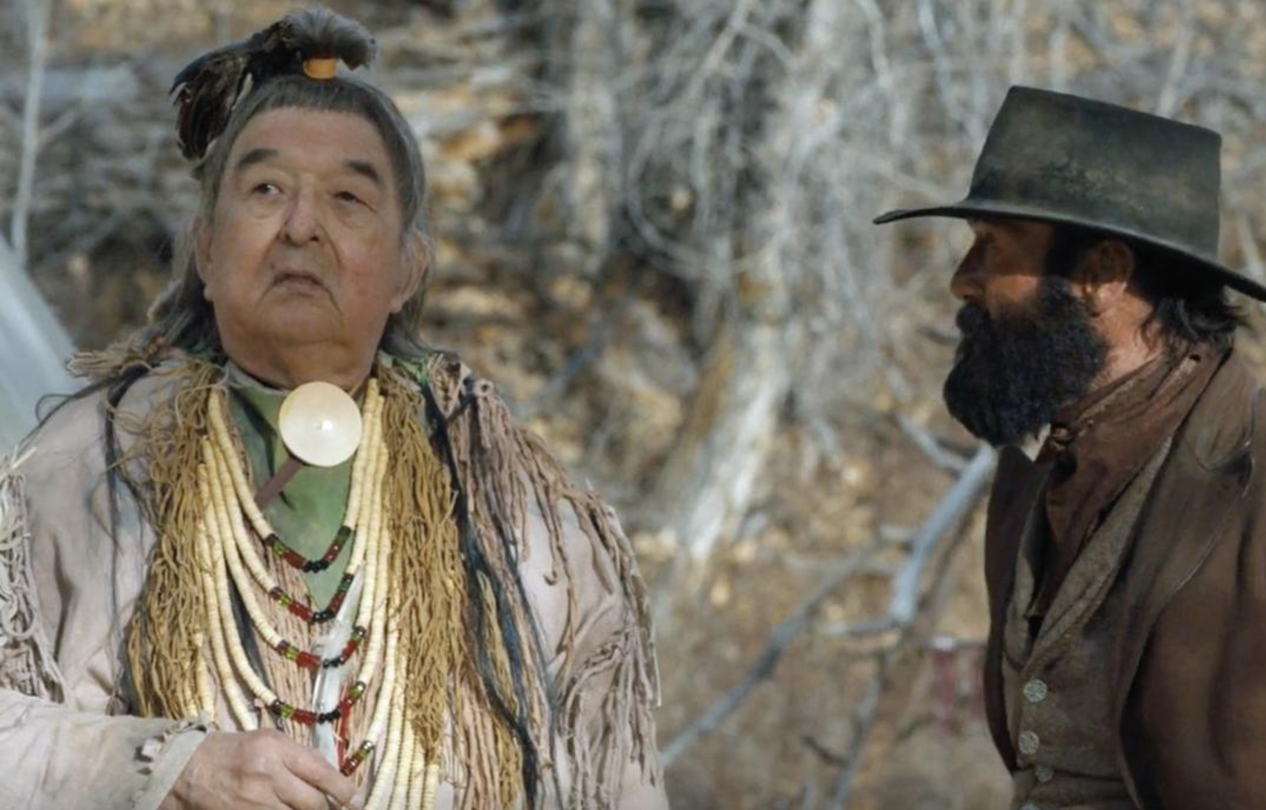 Two men engaged in conversation in a wilderness setting, one dressed traditionally and the other in period clothing.