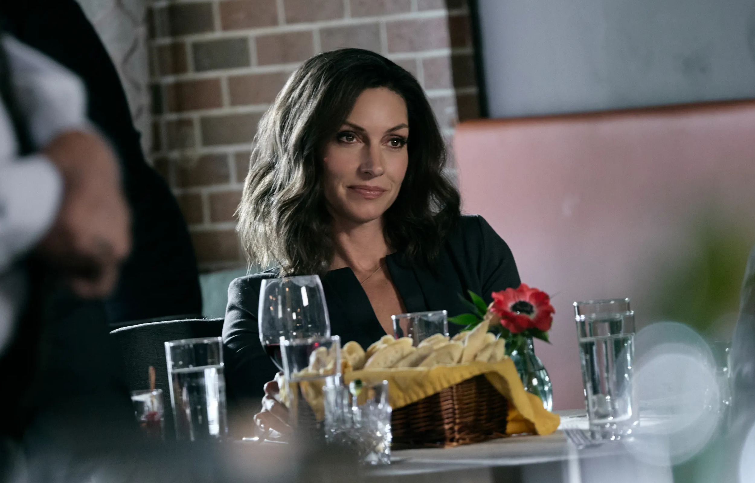 A woman with wavy hair seated at a dining table, looking thoughtfully at the camera with food and drinks in front of her.