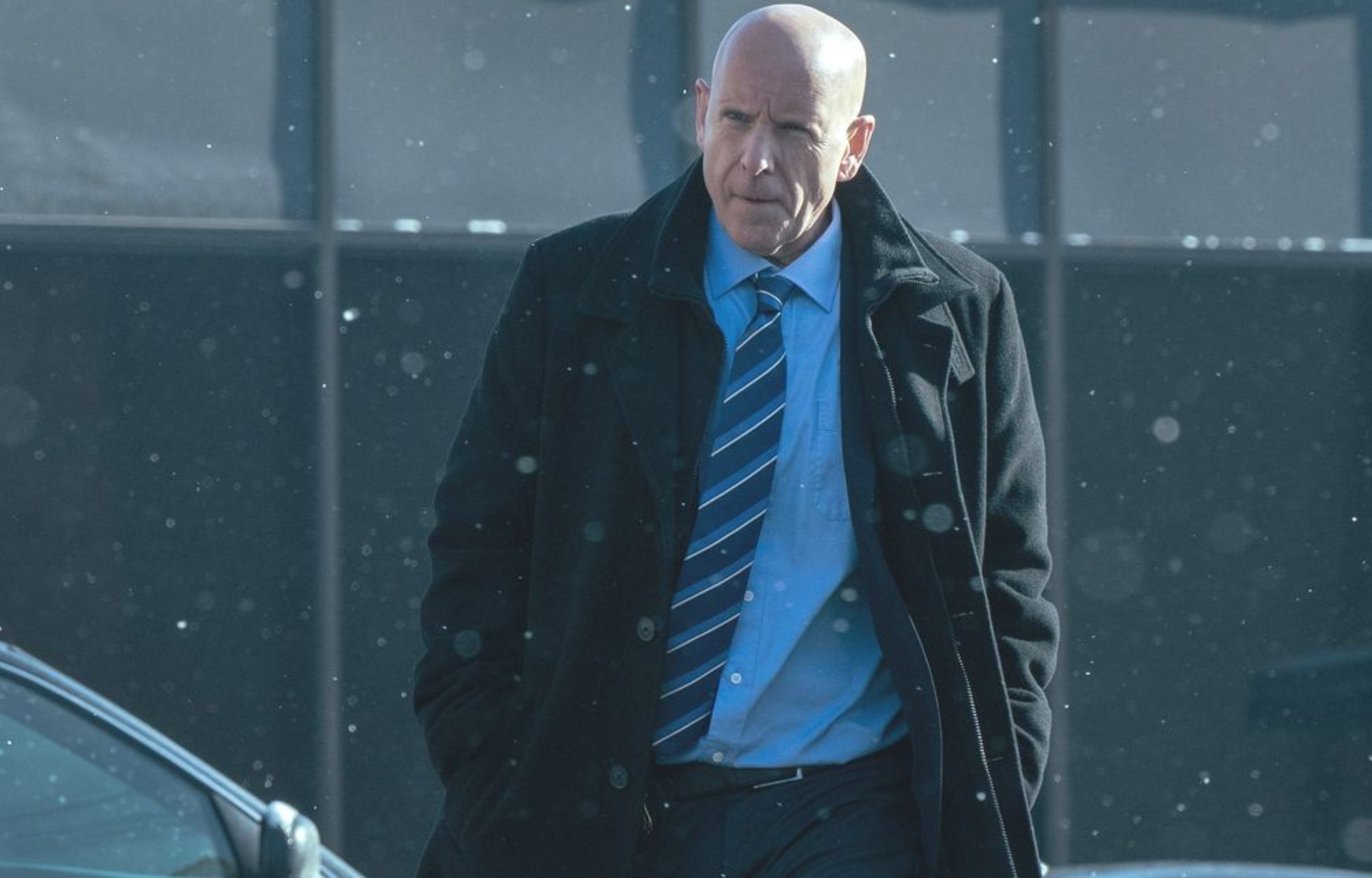 A bald man in a black coat and blue shirt walks outside during snowfall.