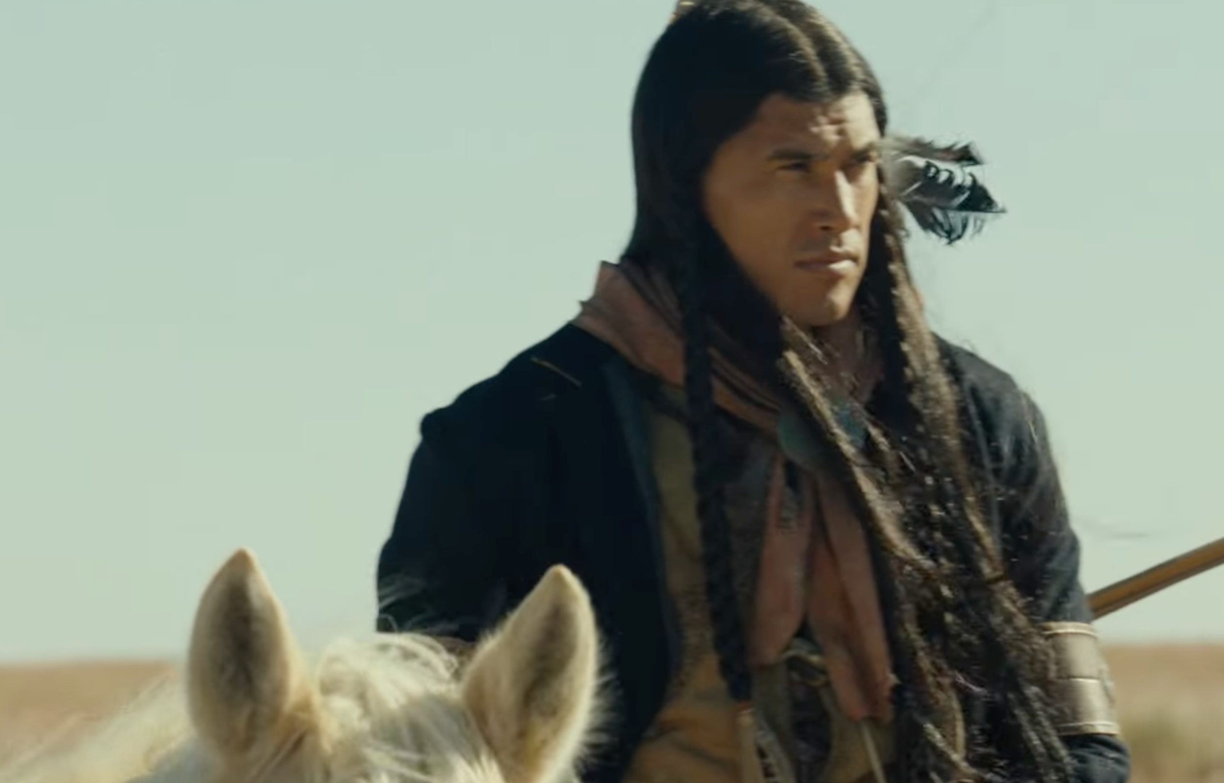 A man with long hair and traditional dress sits on a horse in a vast landscape, gazing into the distance.