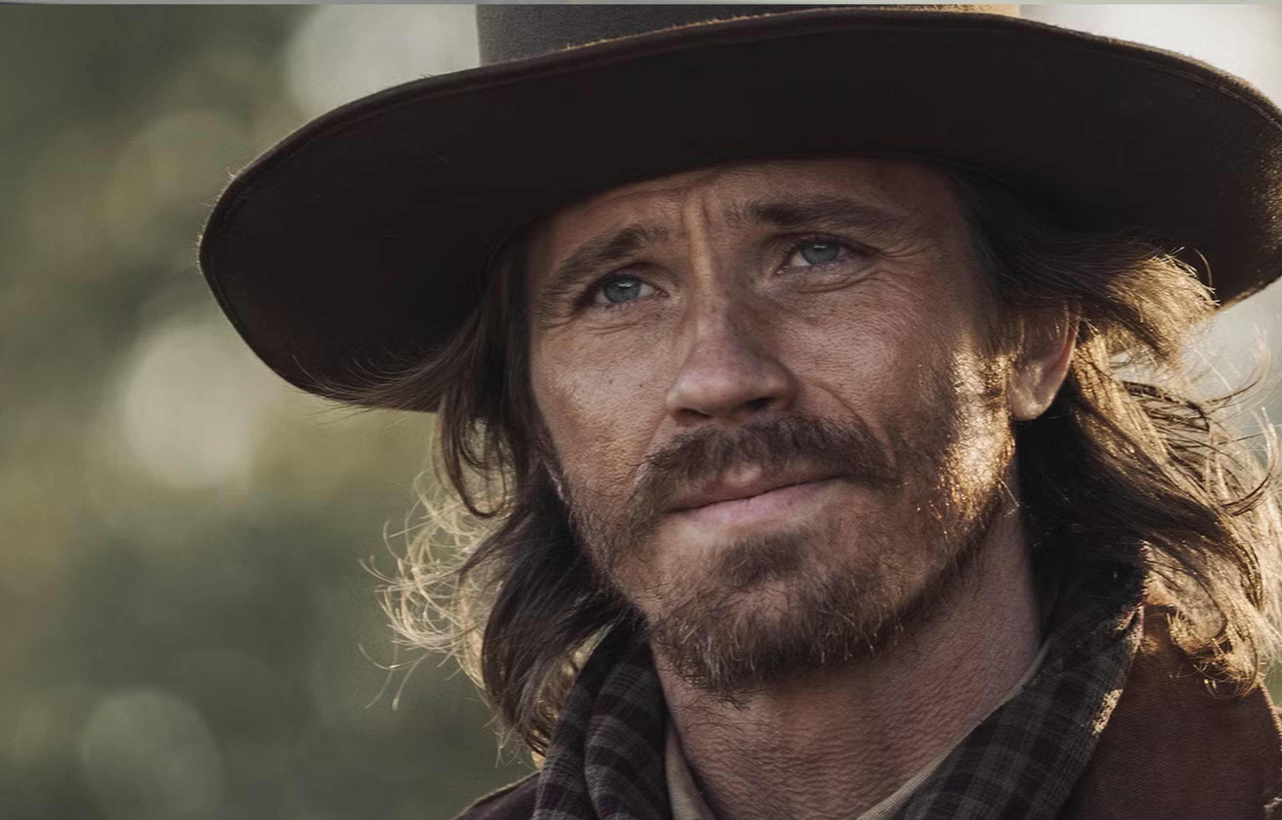 Close-up portrait of a man wearing a wide-brimmed hat, with long hair and a thoughtful expression against a blurred background.