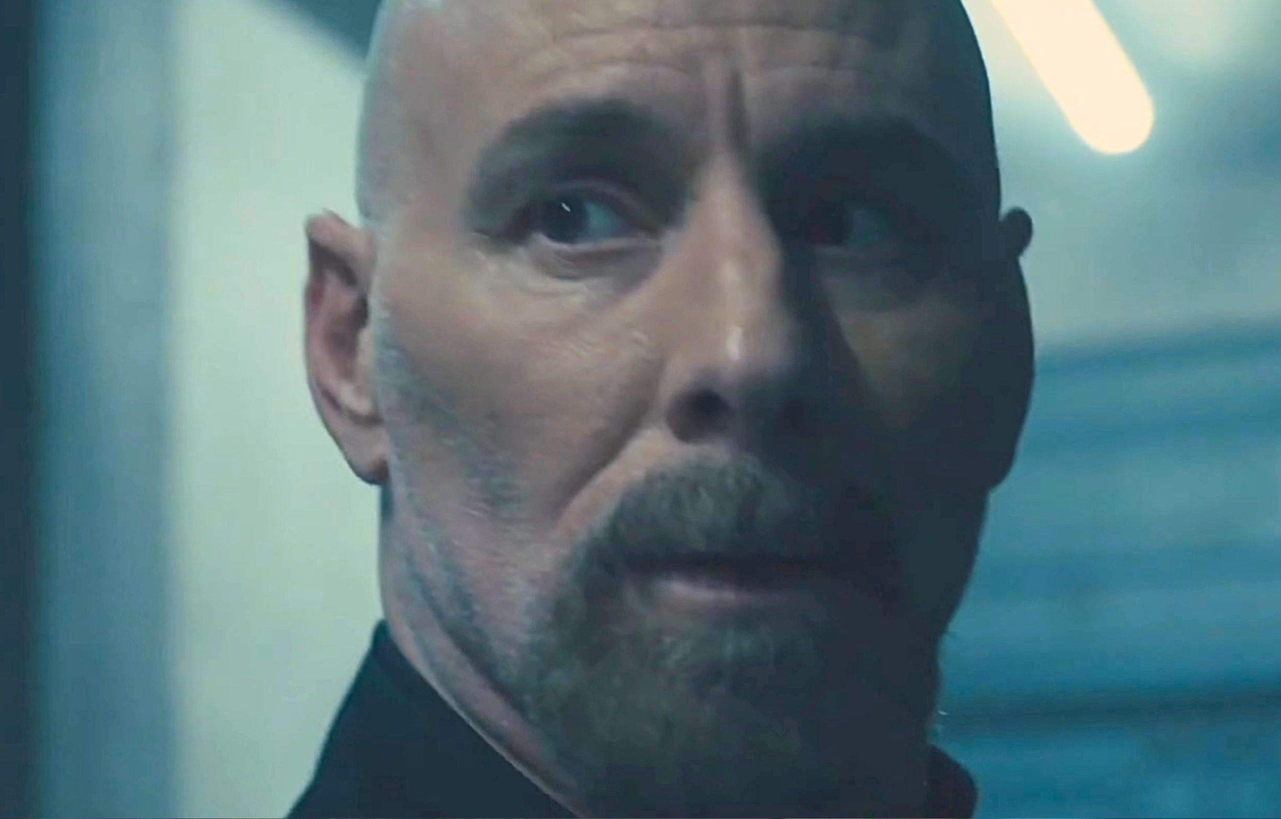 A man with a shaved head and beard looking thoughtfully over his shoulder in a dimly lit environment.