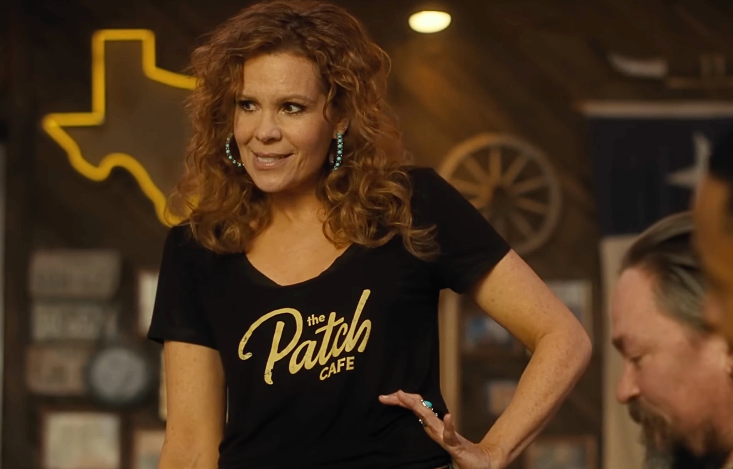 Woman with curly red hair wearing a black shirt with "the Patch Café" logo, smiling in a lively café setting.