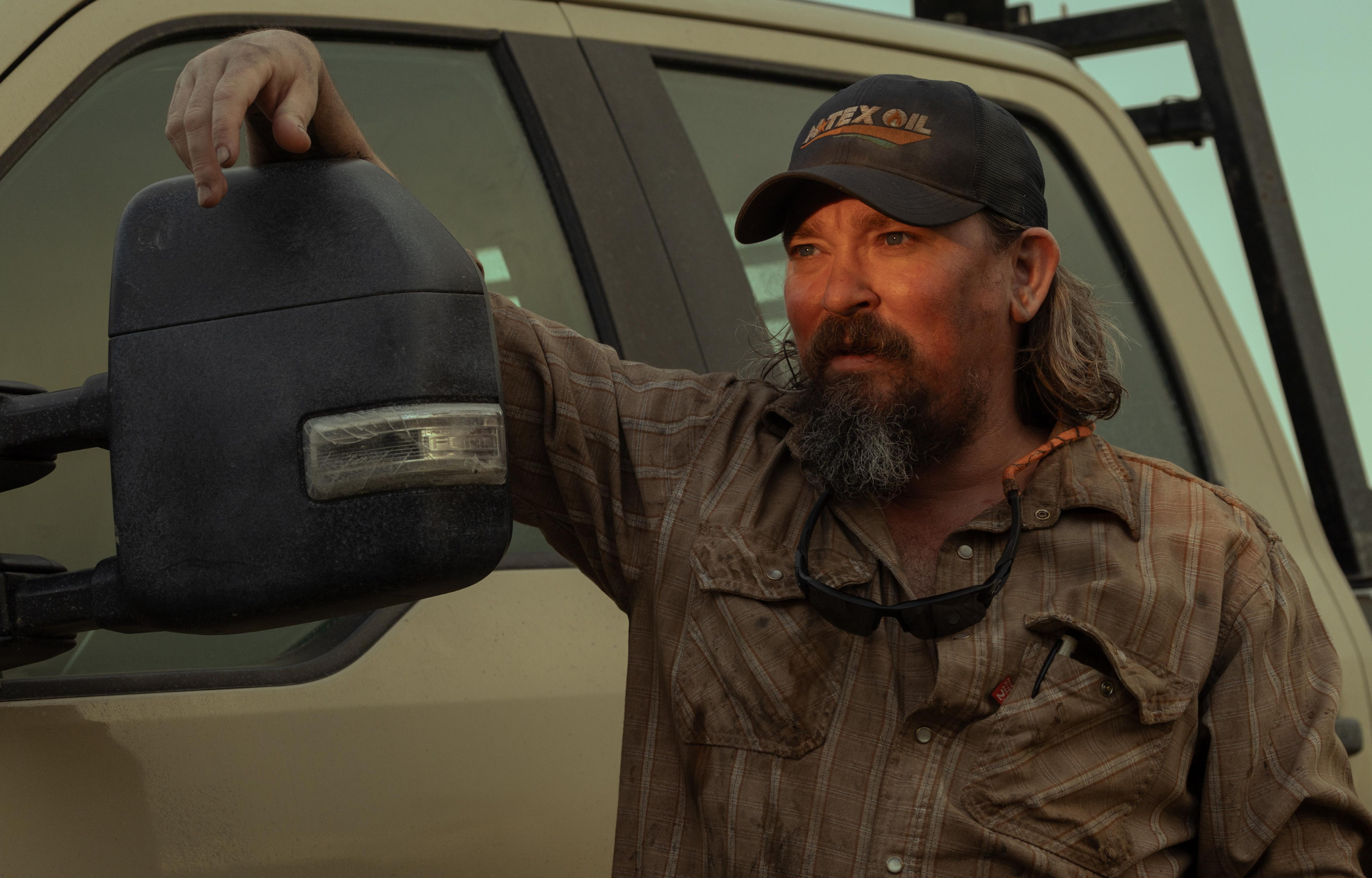 Man with a beard resting his hand on a truck mirror, wearing a plaid shirt and baseball cap, with sunglasses around his neck.