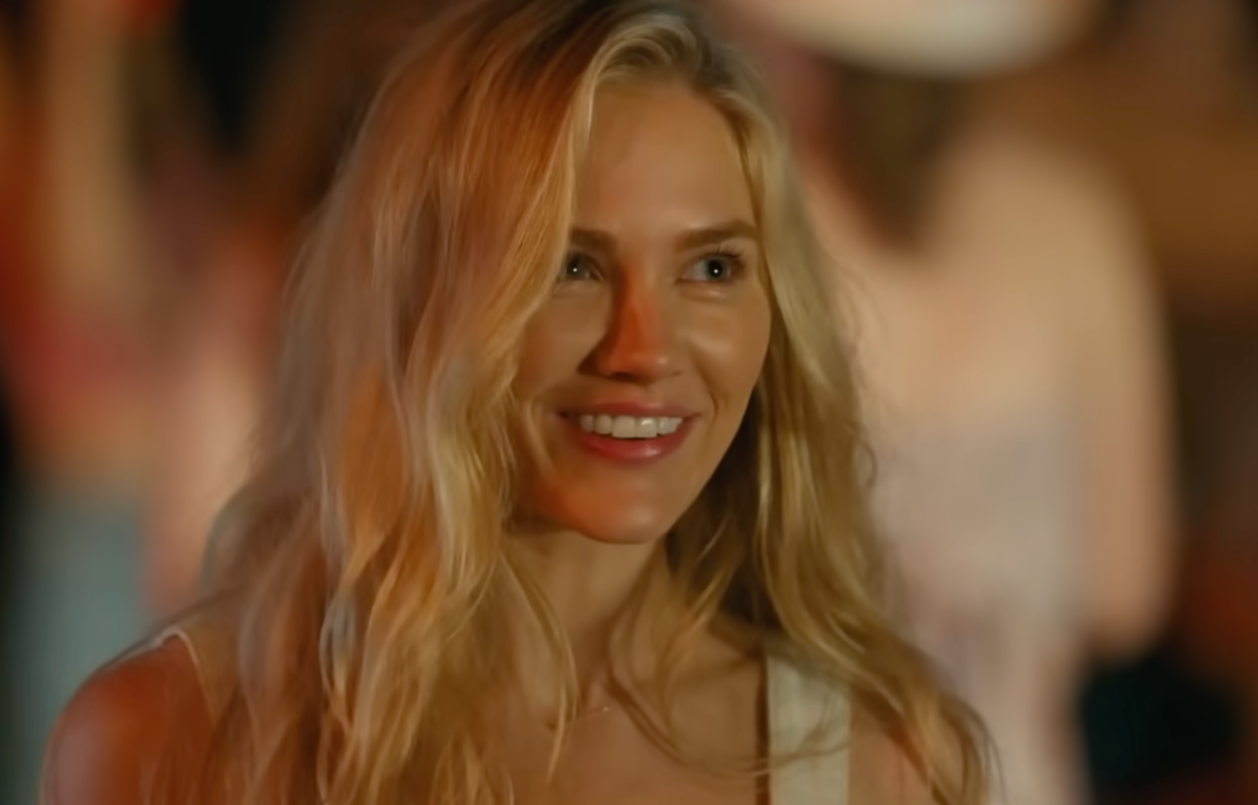 Smiling woman with long, wavy blonde hair, against a blurry background of a lively gathering.